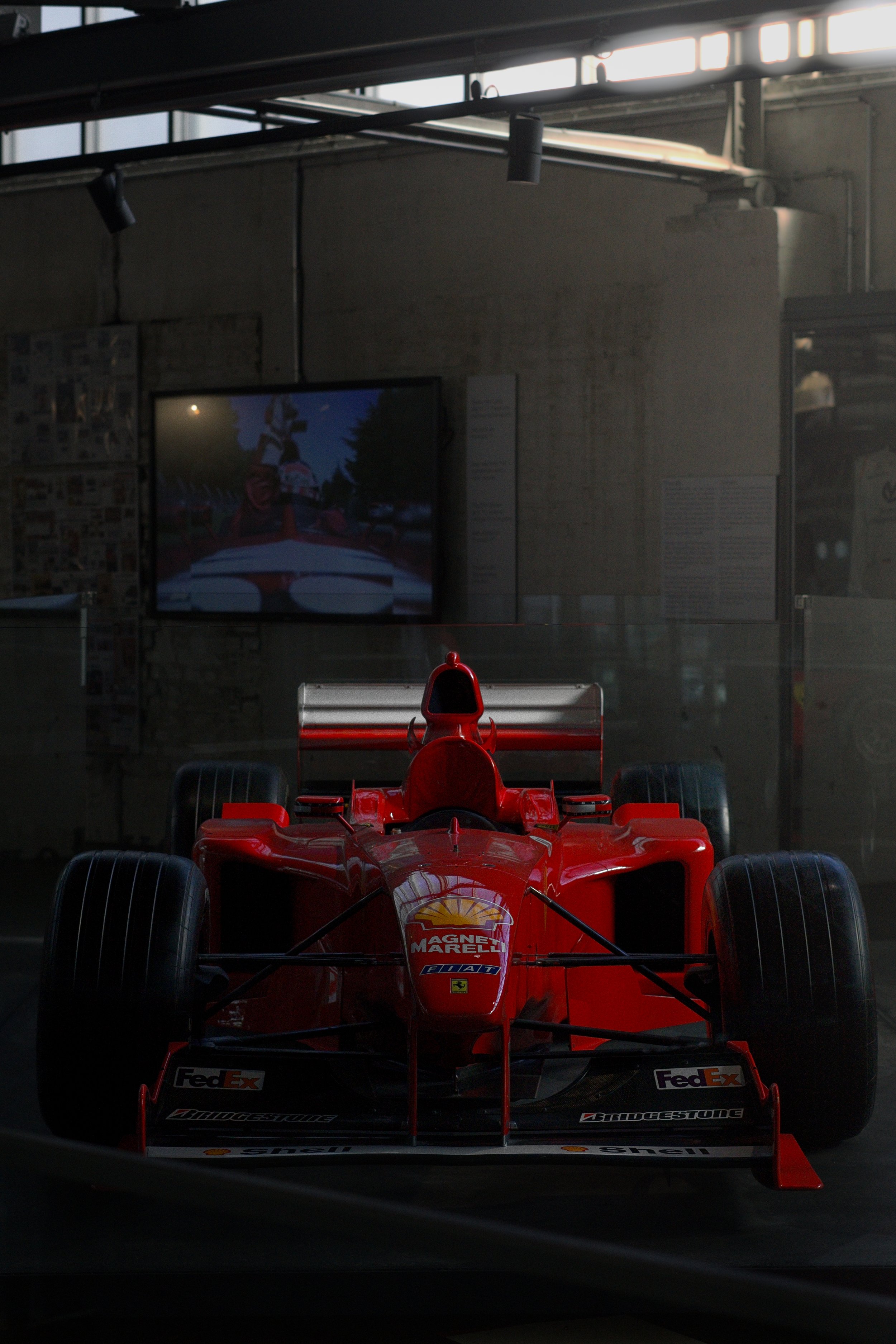 Rotes Formel-1-Auto in einem Museum, im Hintergrund ein Bildschirm mit einem Motorsport-Bild.