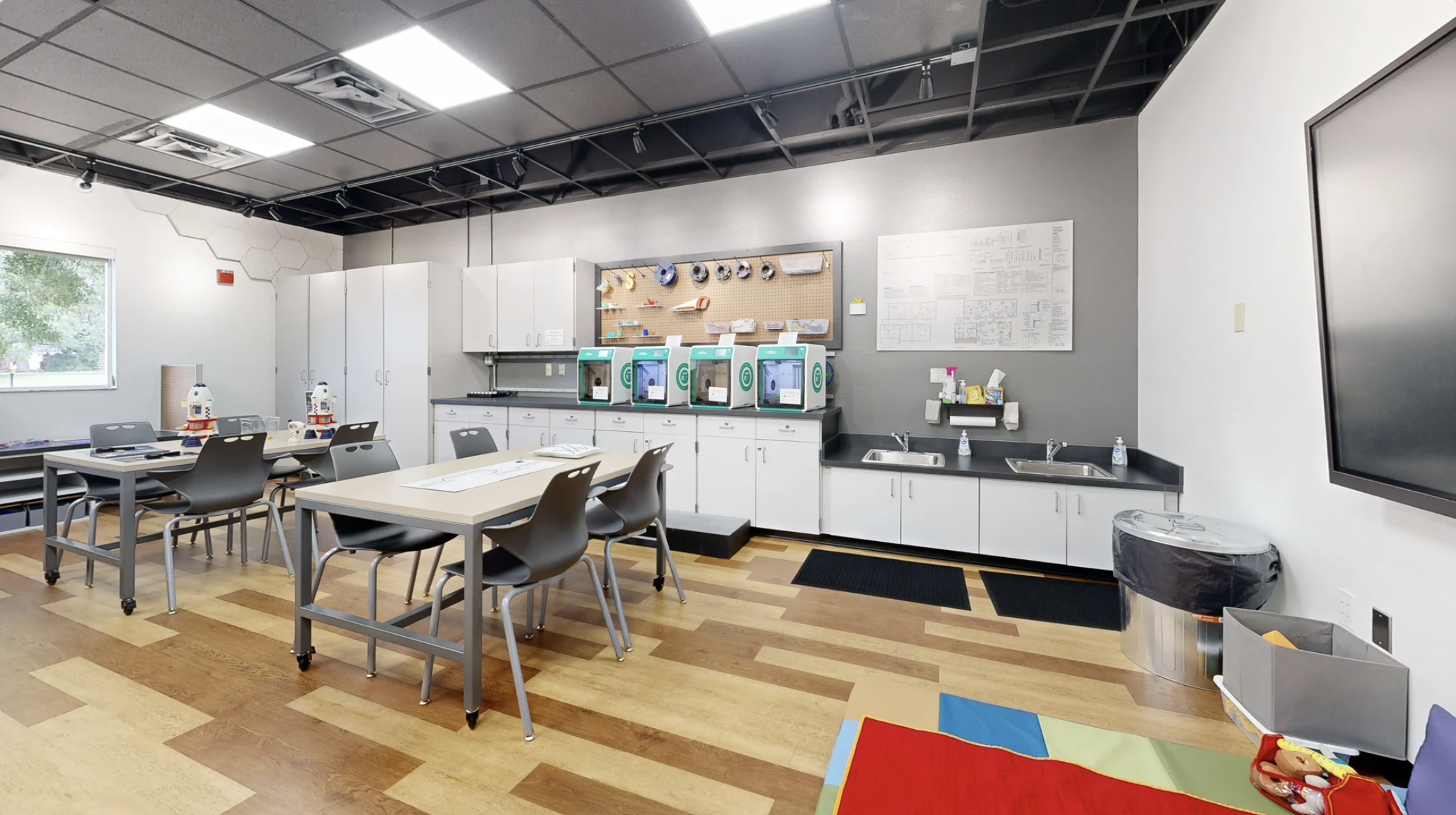 A classroom or workshop room with tables, chairs, and a kitchenette area with sinks, cabinets, and 3 3D printers on the counter. There is a wall-mounted whiteboard and a pegboard with tools or models. The room has a wood floor, gray walls, and a wind