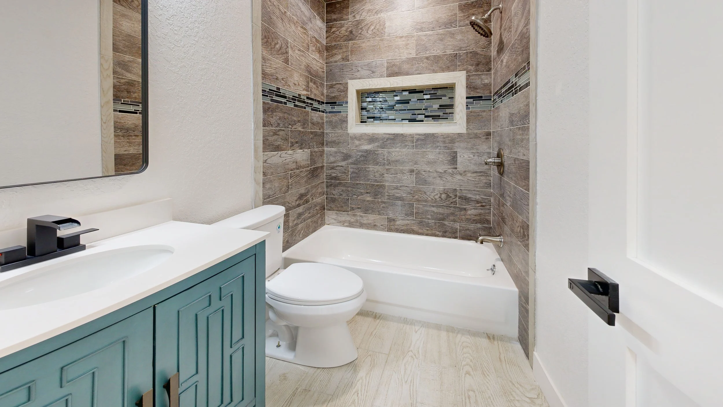 Bathroom with a bathtub, a toilet, a vanity with a sink, a mirror, and a tiled wall with horizontal wood-like tiles and a decorative glass tile strip, and a small window.