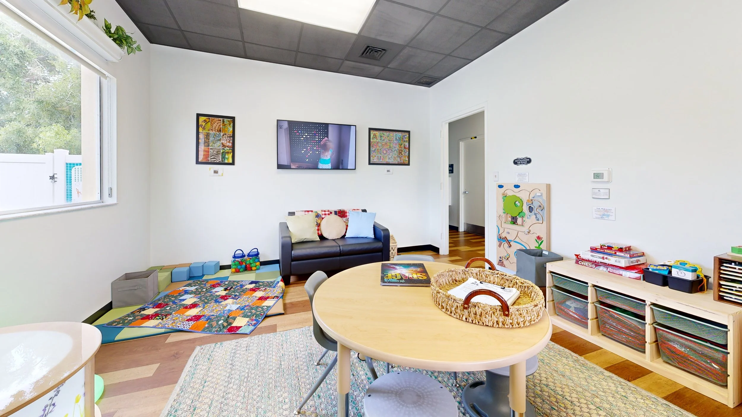 A children's playroom with a round table and chairs, a black couch with colorful pillows, framed artwork on the white walls, and various toys and educational materials on shelves and the floor. A window with a view of trees is visible on the left.
