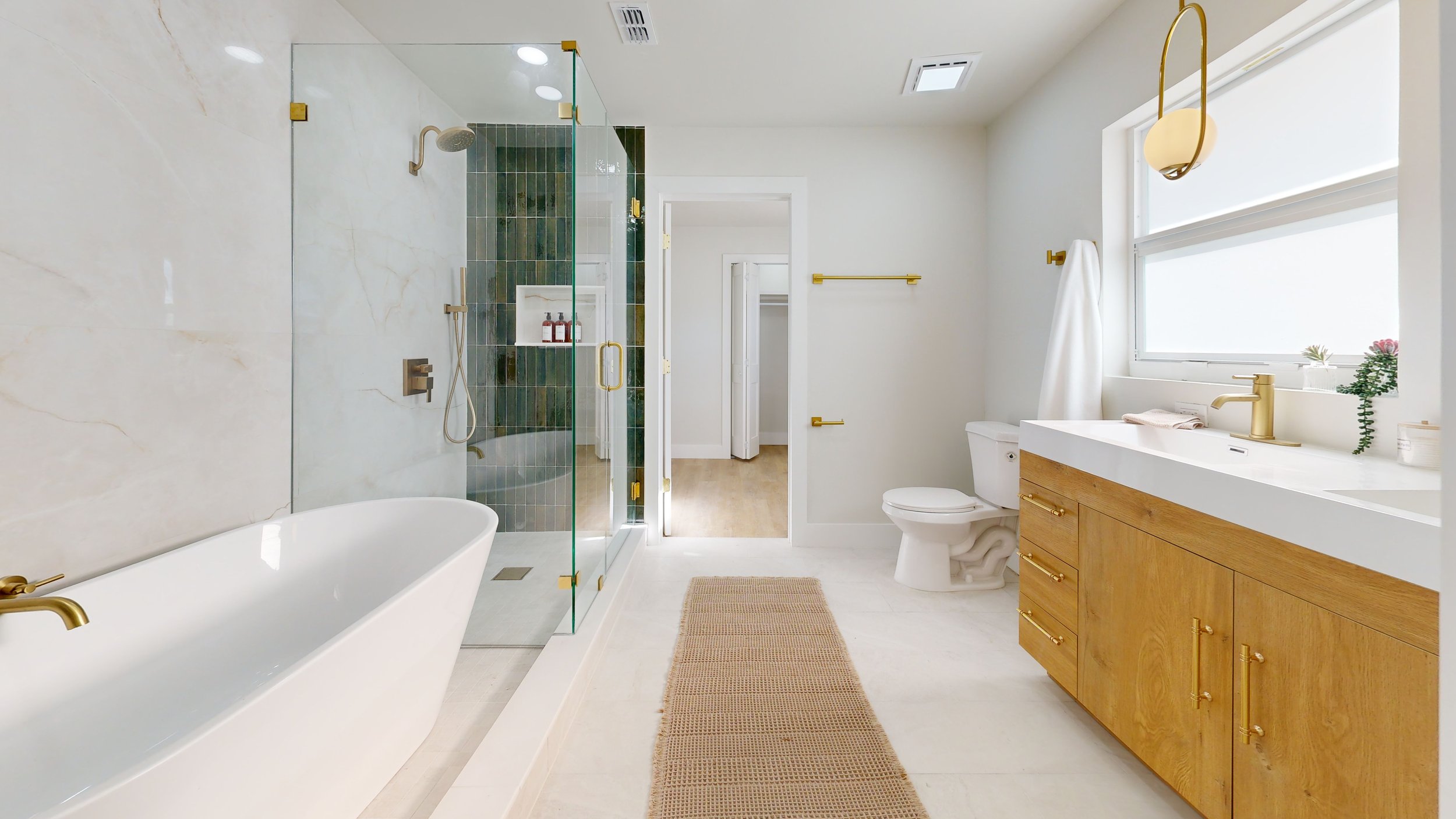 Modern bathroom featuring a bathtub, glass shower enclosure with green tiles, white walls, a wooden vanity with gold hardware, a white toilet, and a large window with white curtains, decorated with plants.