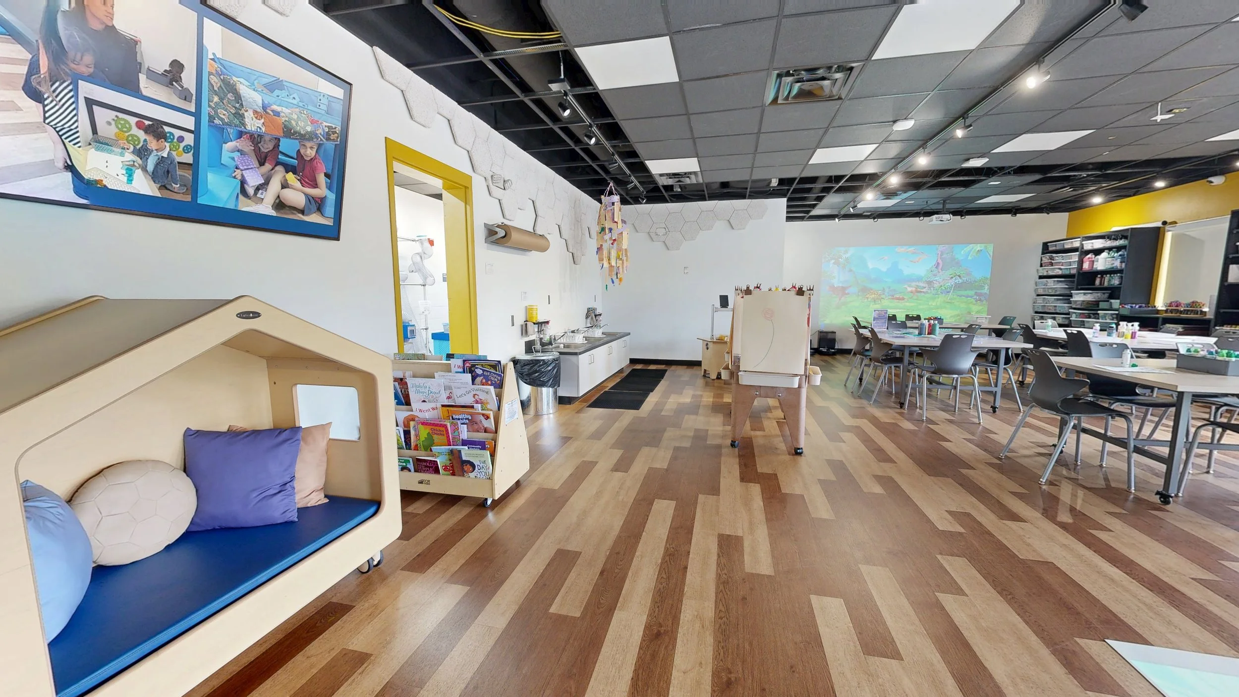 A spacious and colorful children's activity and learning space with tables, shelves, a reading nook with pillows, and various art supplies, with a digital projector displaying a landscape scene on the back wall.