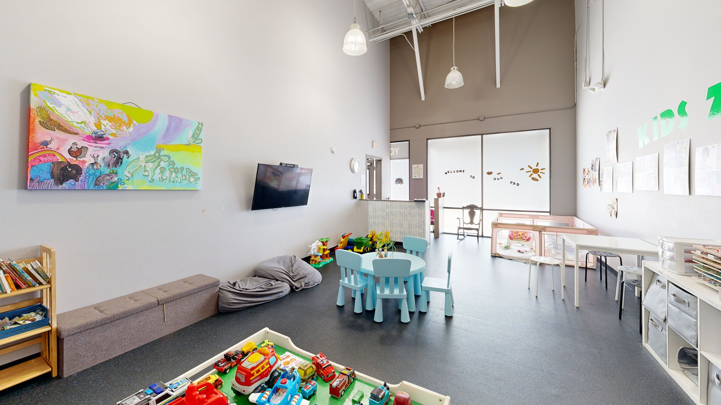 A spacious indoor children's play area with white walls, colorful artwork, toys, small tables, and chairs. There is a television mounted on the wall and a window allowing natural light. The space is decorated for children with playful and inviting fu