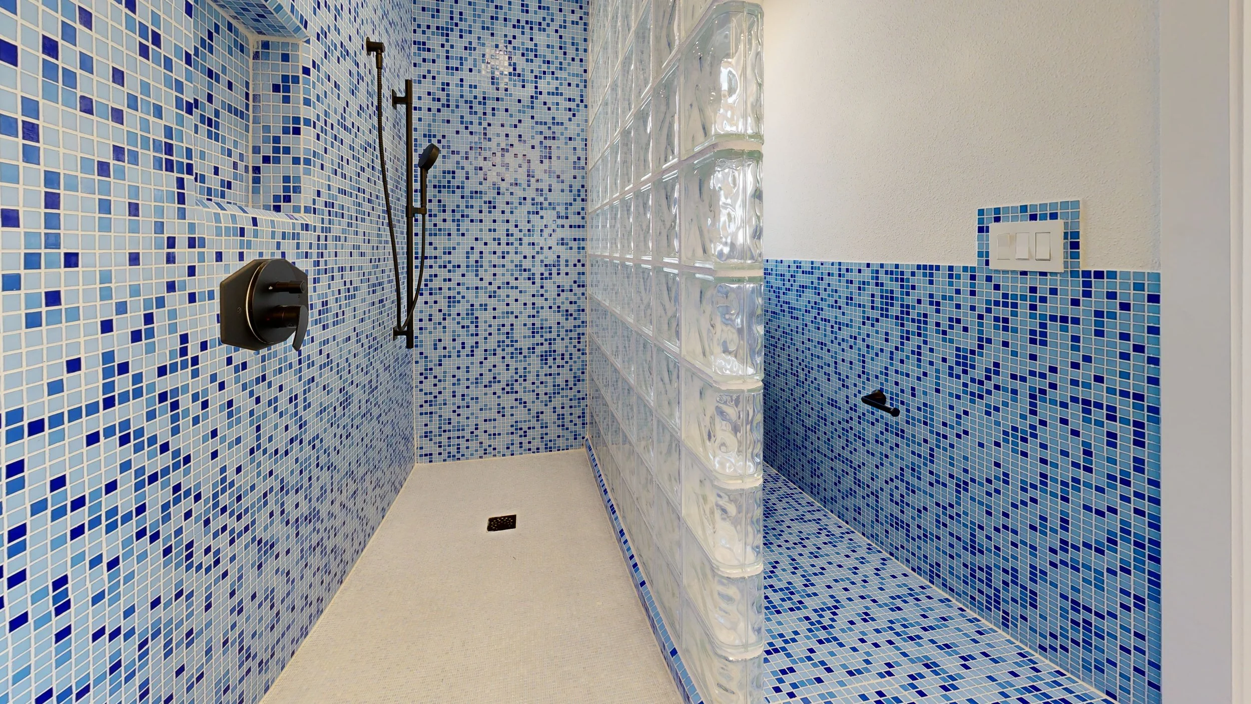 A shower area with walls covered in blue and white mosaic tiles, a glass block partial wall, and a separate shower head with controls.