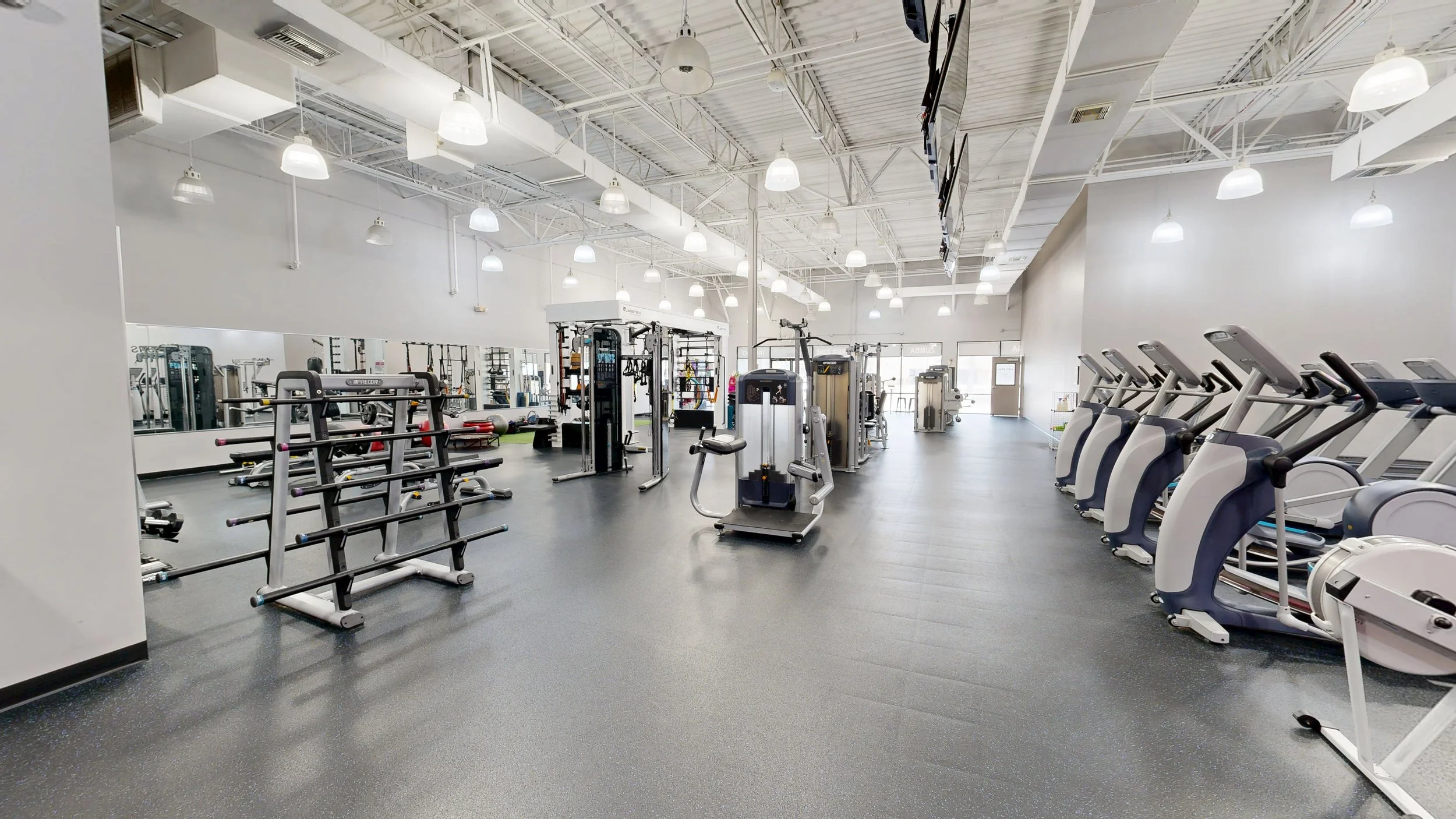Empty gym with various workout equipment including treadmills, ellipticals, weight machines, and free weights, with bright overhead lighting and large mirrors on the wall.