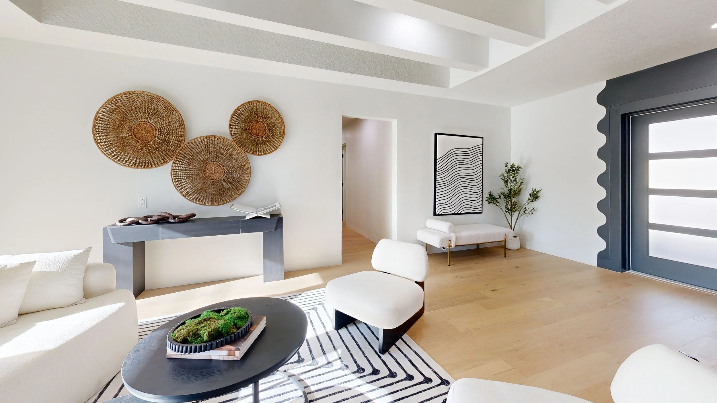 Modern living room with white walls, wooden flooring, a black striped rug, and contemporary furniture including white chairs and a beige sofa. Decor includes woven wall art, a black and white framed art piece, a plant, and a black coffee table with a garden tray on top. There's a large gray door with a scalloped edge and a window with a wavy border.