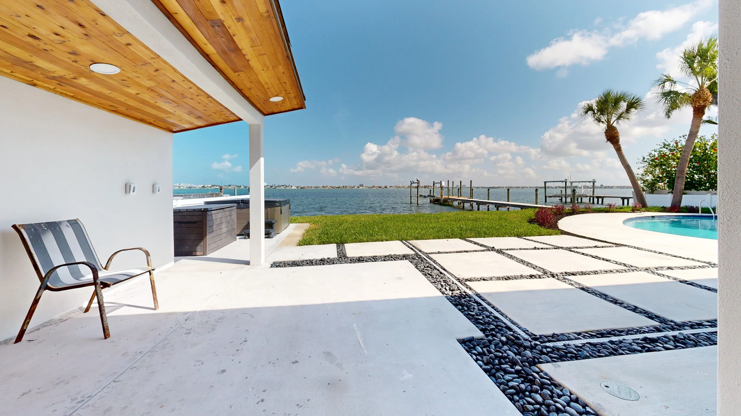 A patio overlooking a waterway with a hot tub, palm trees, and a boat dock under a blue sky with scattered clouds.