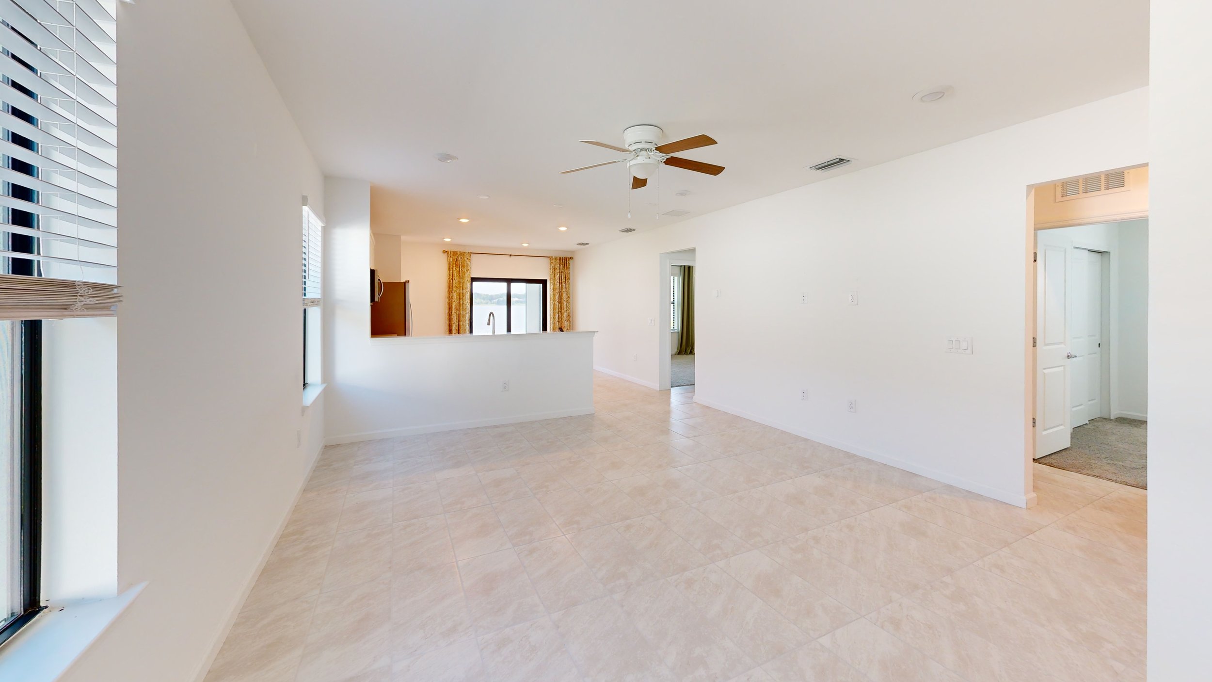 Empty living room with tiled floor, white walls, ceiling fan, window with blinds, and sliding door leading outside.