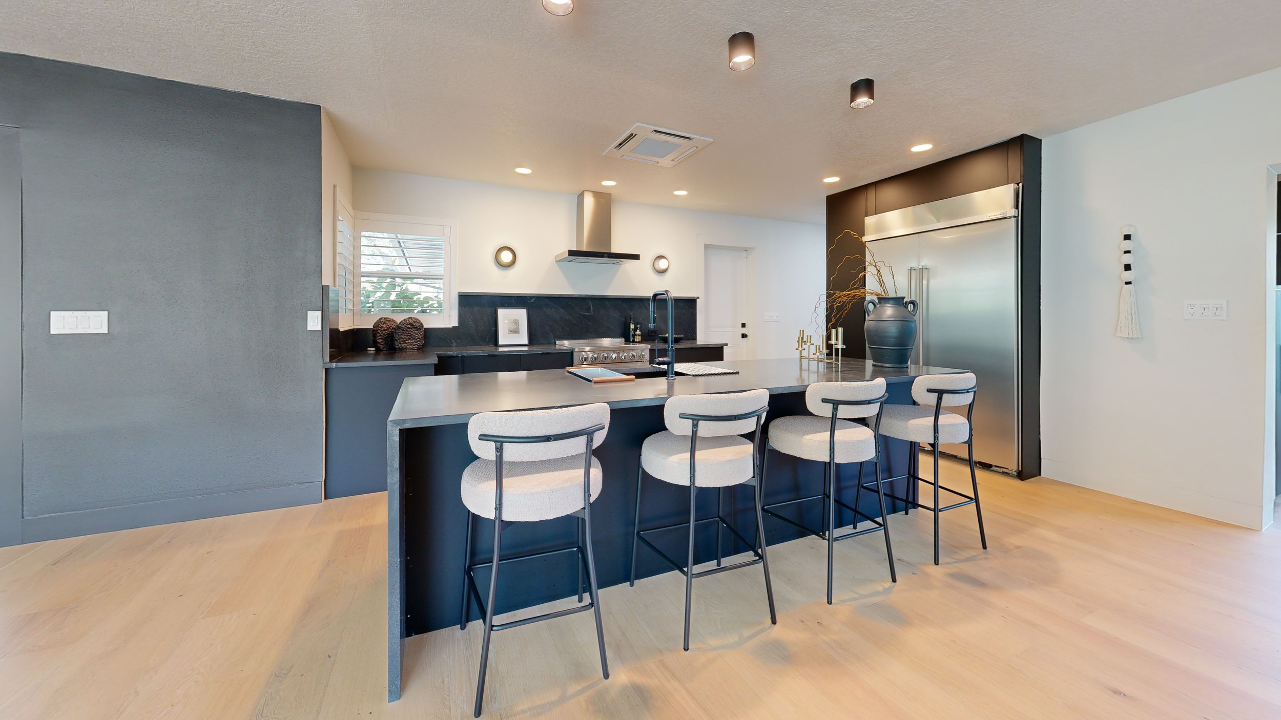Modern kitchen with a black and white color scheme, featuring a kitchen island with four upholstered chairs, black countertop, stove, stainless steel refrigerator, black and gold decorative items, and a window with plantation shutters.