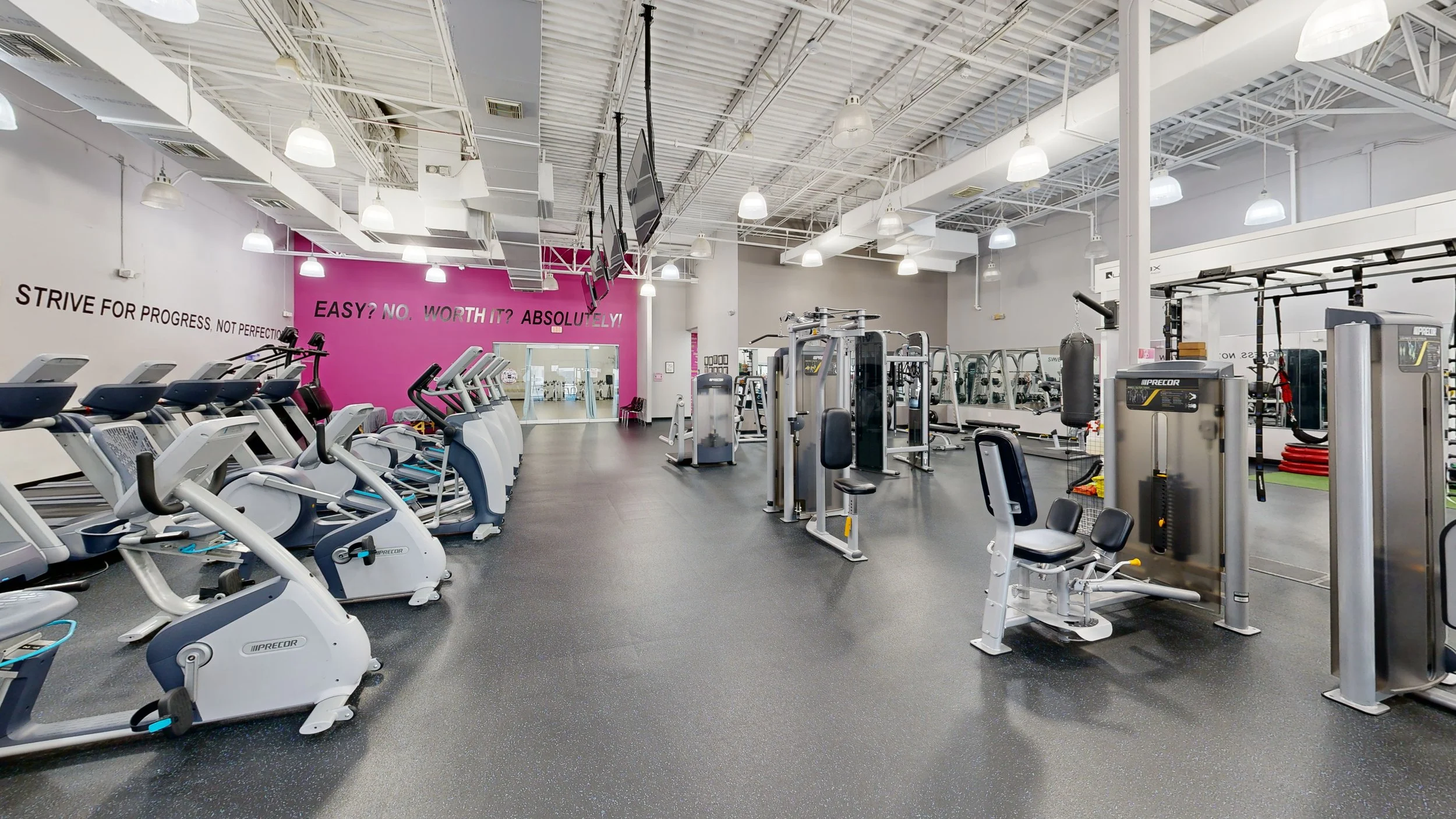 Interior of a modern gym with various exercise equipment including treadmills, ellipticals, weight machines, and punching bag. Bright lighting and motivational wall messages.
