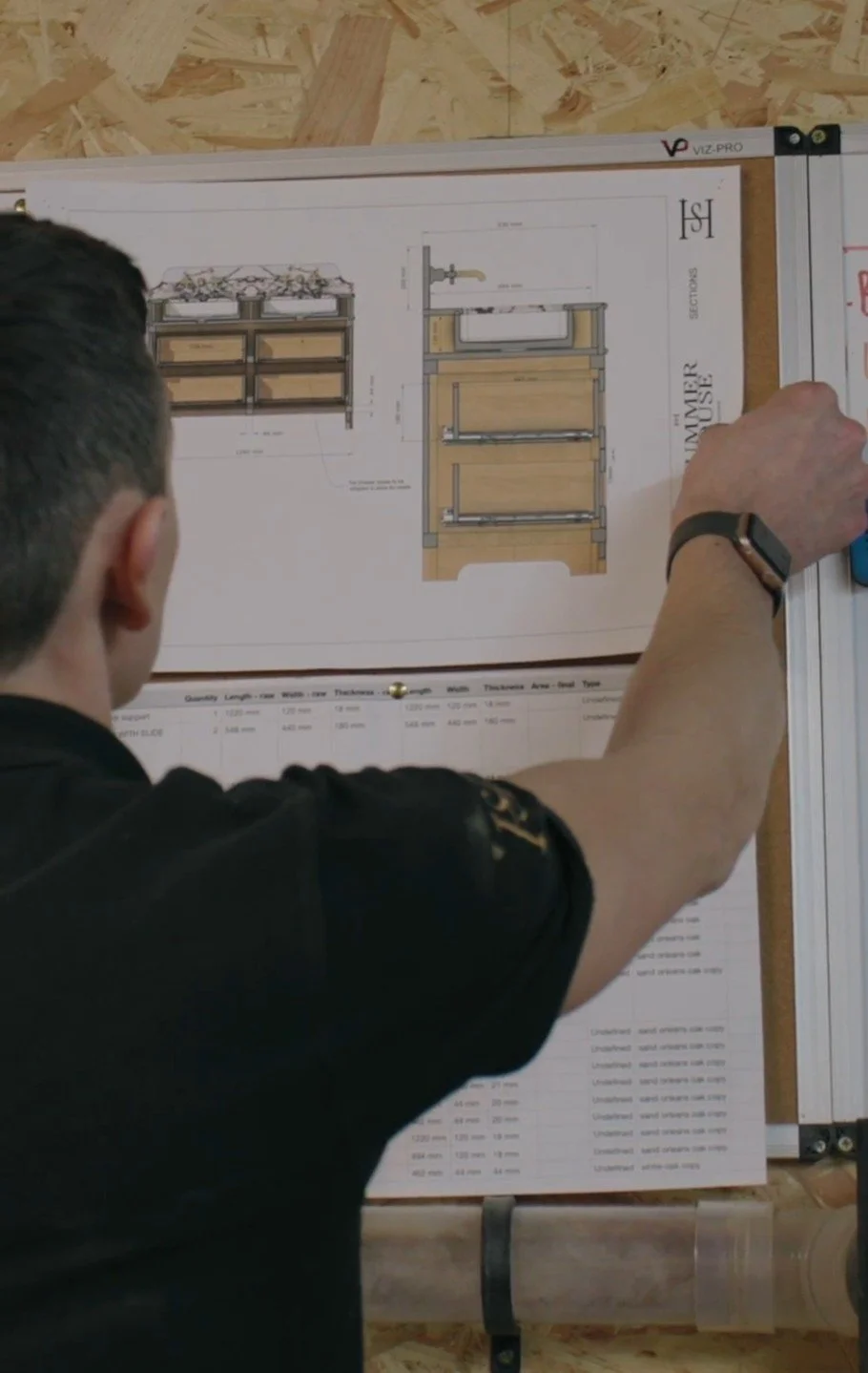 A person with short dark hair and wearing a black shirt is pointing at a detailed architectural design for a kitchen sink and cabinet layout on a board.