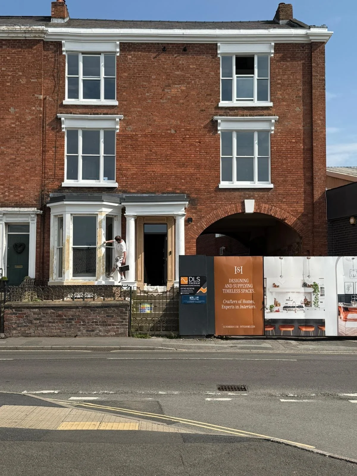 A man working on the front porch of a three-story brick house under renovation. The house has four large white-framed windows, and there is a construction sign in front advertising interior design services.