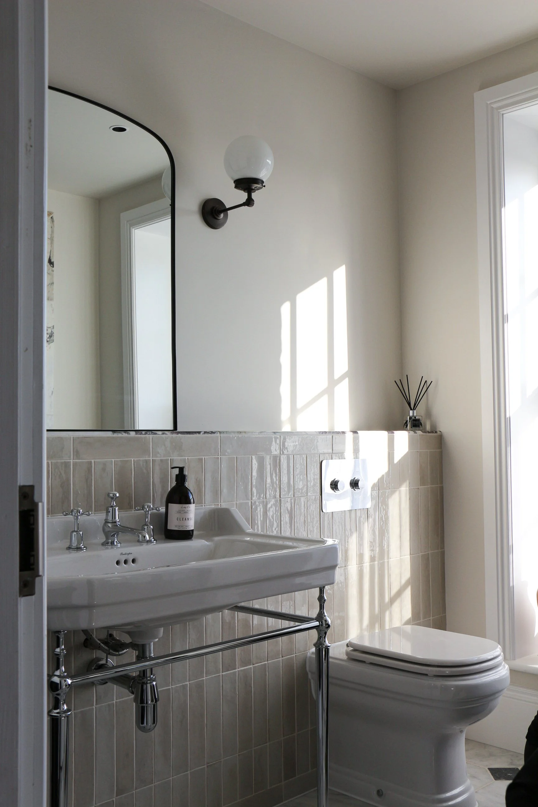 A bathroom with a white porcelain sink and a toilet. The sink has a black soap dispenser and vintage-style chrome faucets. Above the sink is a mirror and a wall-mounted light fixture with a globe-shaped bulb. Natural sunlight creates shadow patterns on the wall.