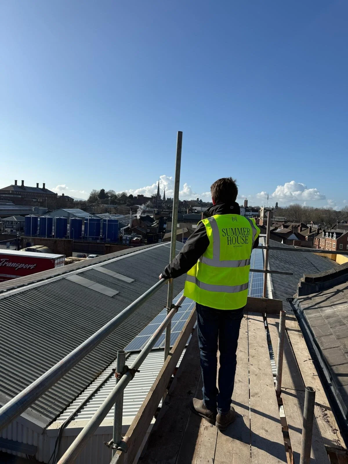 A man in a yellow vest labeled 'Summer House Interiors' standing on scaffolding on a rooftop with solar panels, overlooking a cityscape with buildings, rooftops, and a blue sky with some clouds.