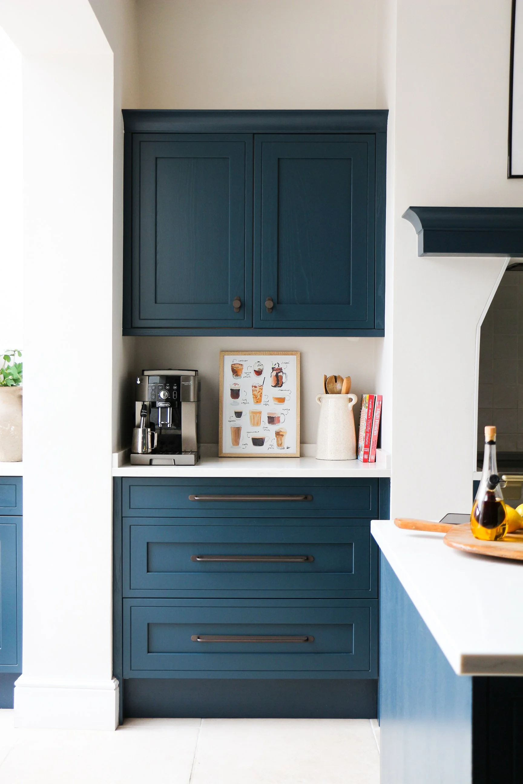 Blue kitchen cabinets with silver handles in a modern kitchen, coffee machine, framed coffee chart, and decorative items on a white countertop.