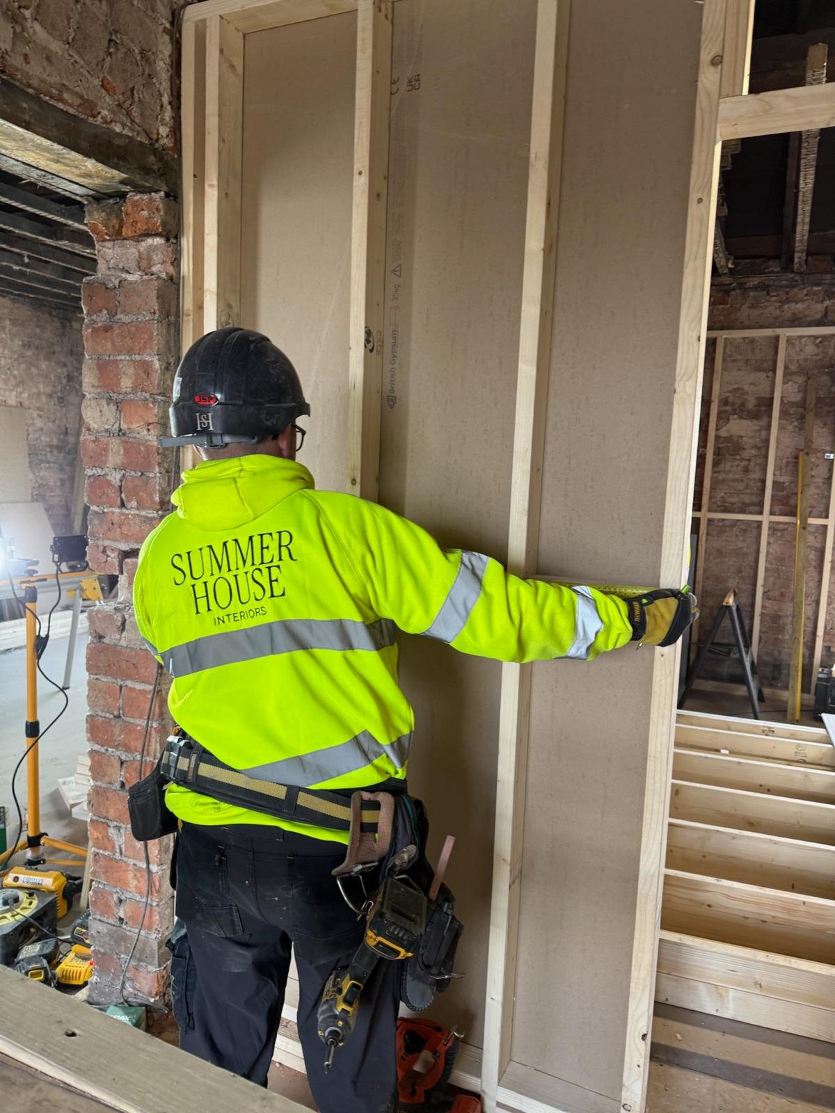 A construction worker wearing a yellow high-visibility jacket with the words 'Summer House Interiors' on the back, a black safety helmet, and safety gloves, working inside a partially built wooden wall frame in a building under renovation.