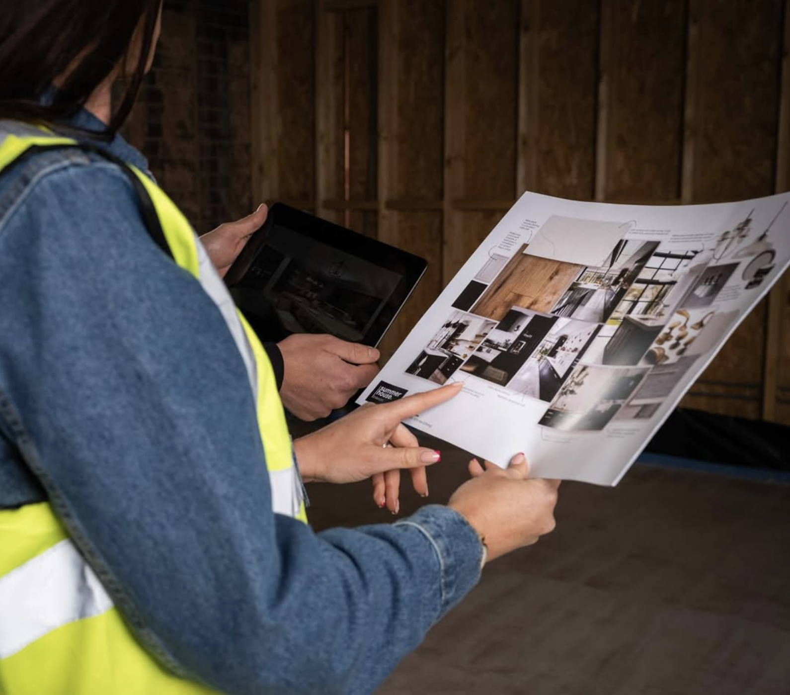 Two people, one with a reflective safety vest and denim jacket, are looking at a printed interior design mood board or brochure, with one person pointing at an image. The background shows a wooden wall.