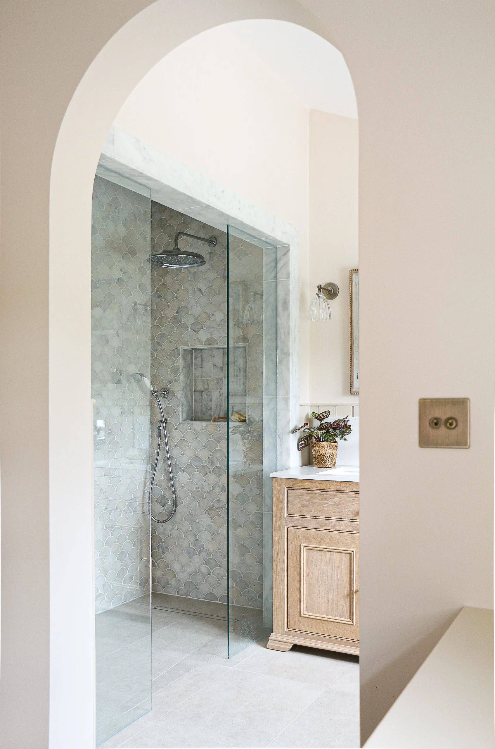 Bathroom with walk-in shower featuring scalloped tile design, a rainfall showerhead, a handheld shower, a window, a wall light, a potted plant on a wooden cabinet, and an arch doorway.