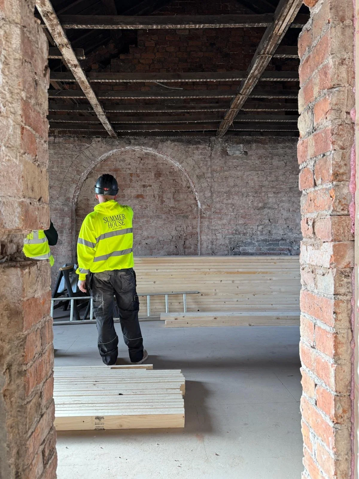 Construction workers in yellow safety jackets and helmets standing inside a brick building under renovation, with stacks of wood and building materials on the floor.