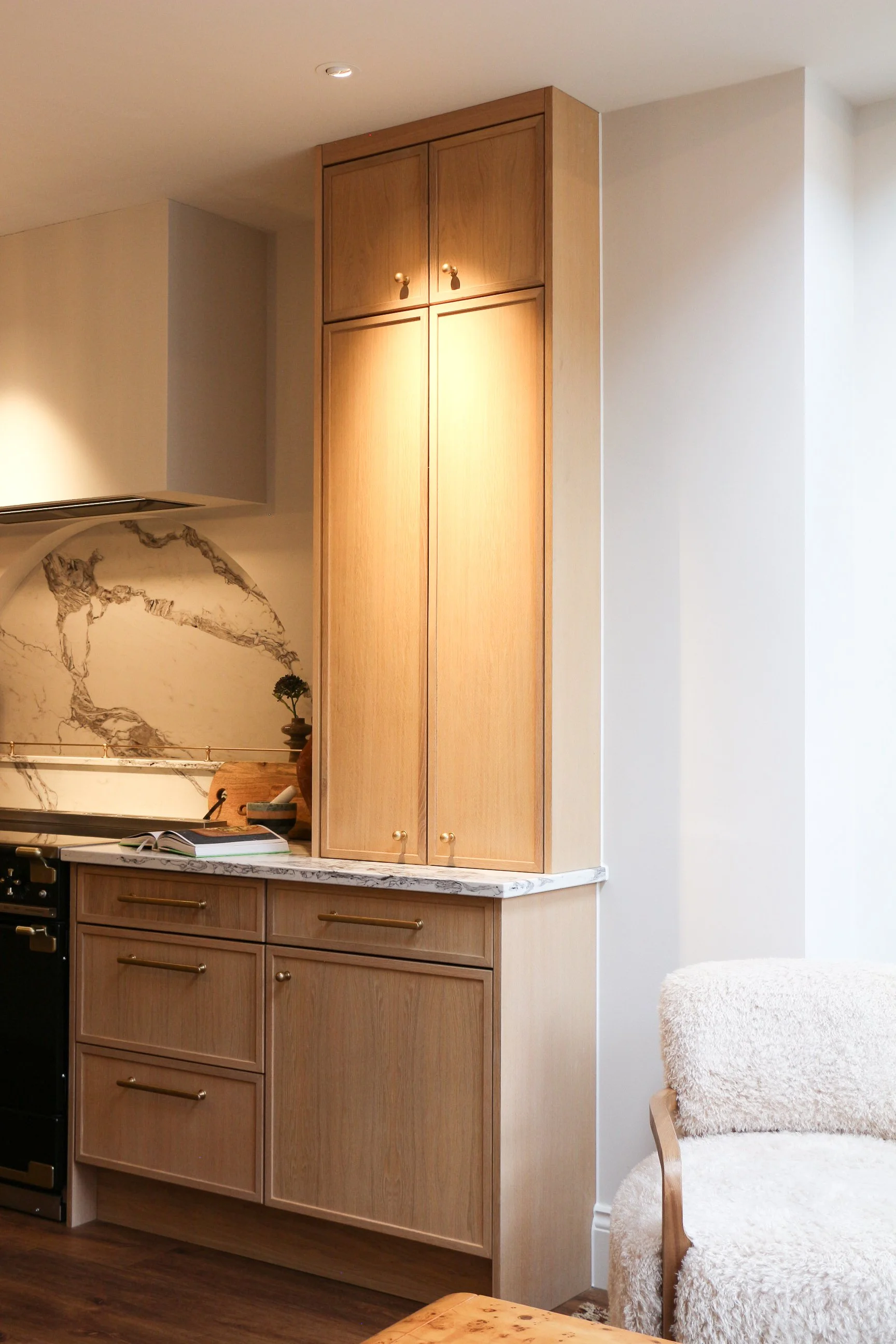 A kitchen corner with beige cabinets, a marble countertop, a small plant, and a black oven. There is a wooden dining table and a chair with a white fluffy cushion next to a light-colored wall.