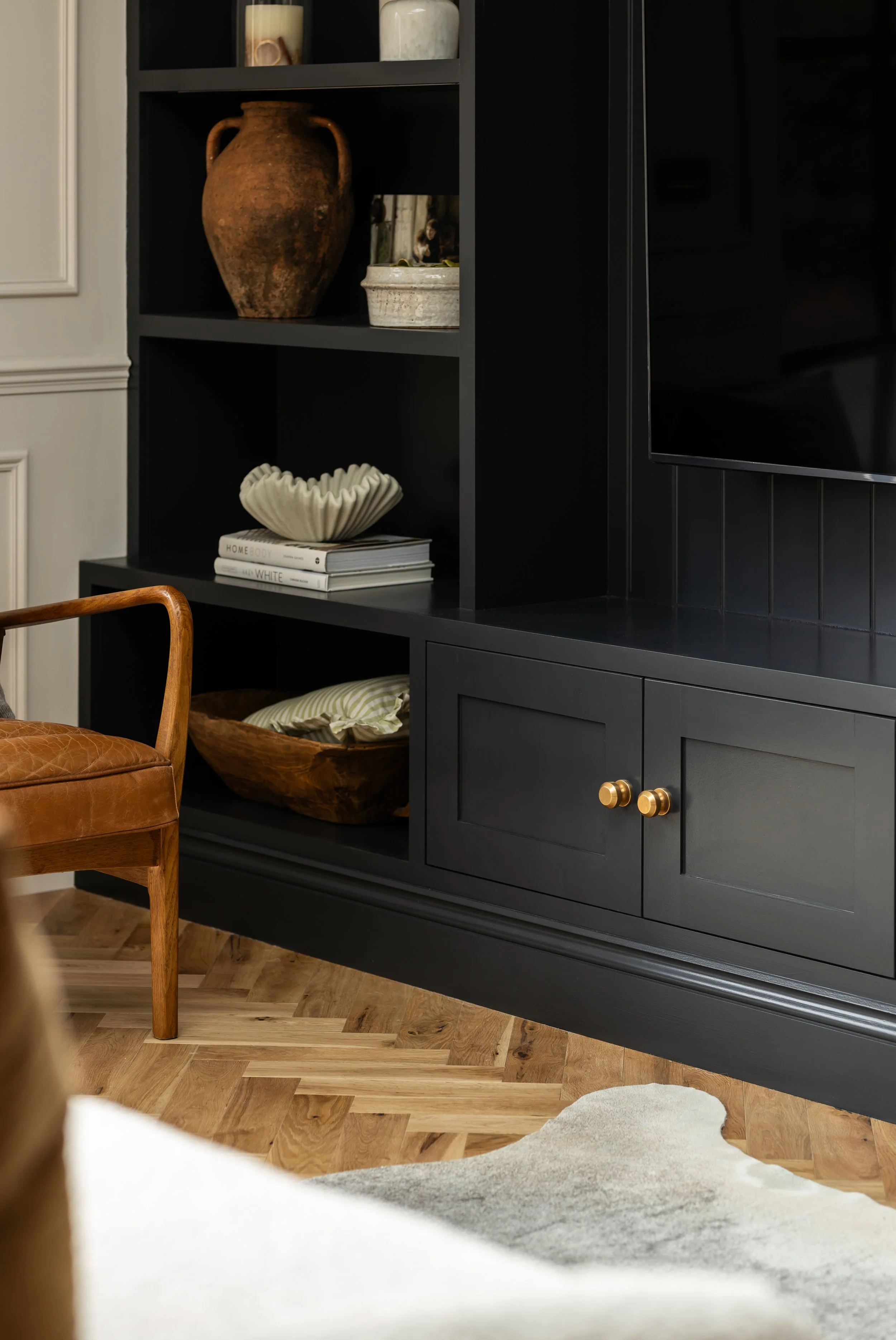 A black bookshelf with decorative items, including a large earthenware jug, candles, a ceramic bowl, a stack of books, and a large ceramic shell sculpture, in a room with a wooden floor and a beige wall.