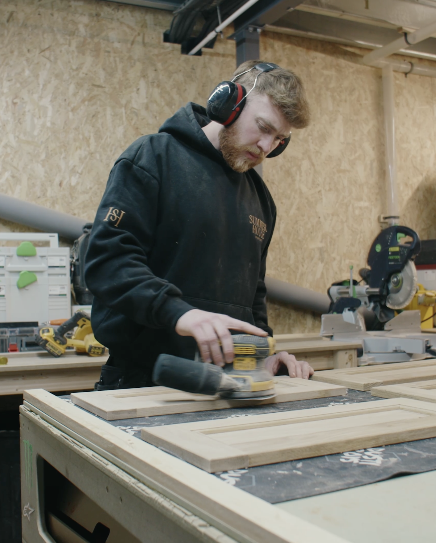 A man working in a woodworking shop, using a power sander on a piece of wood. He is wearing a black hoodie with the text 'SUMMER HOUSE' and ear protection.