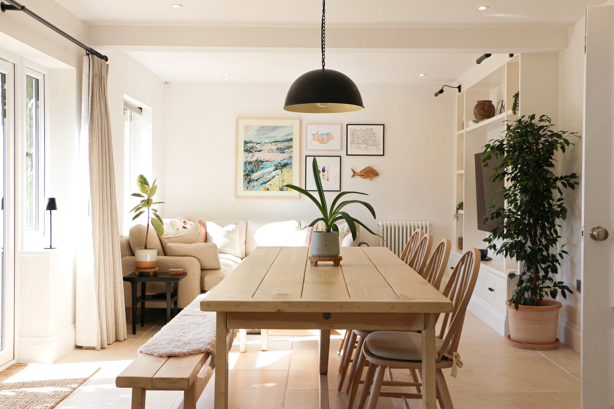Bright living room with a wooden dining table, plants, framed artwork, and a beige sofa.