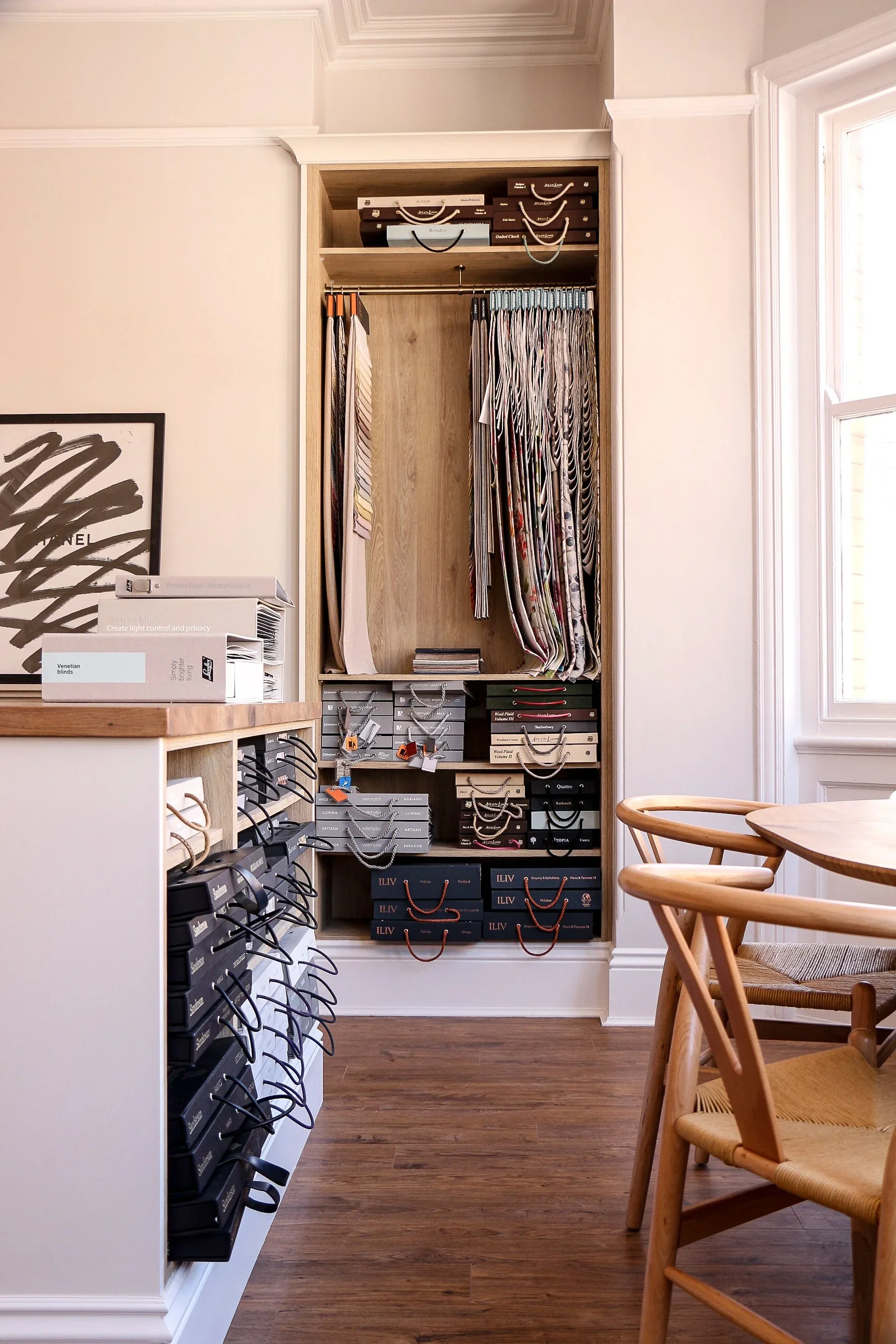 A display of sample necklaces hanging on hooks inside a built-in wooden cabinet in a room with hardwood flooring, surrounded by chairs and a large window.