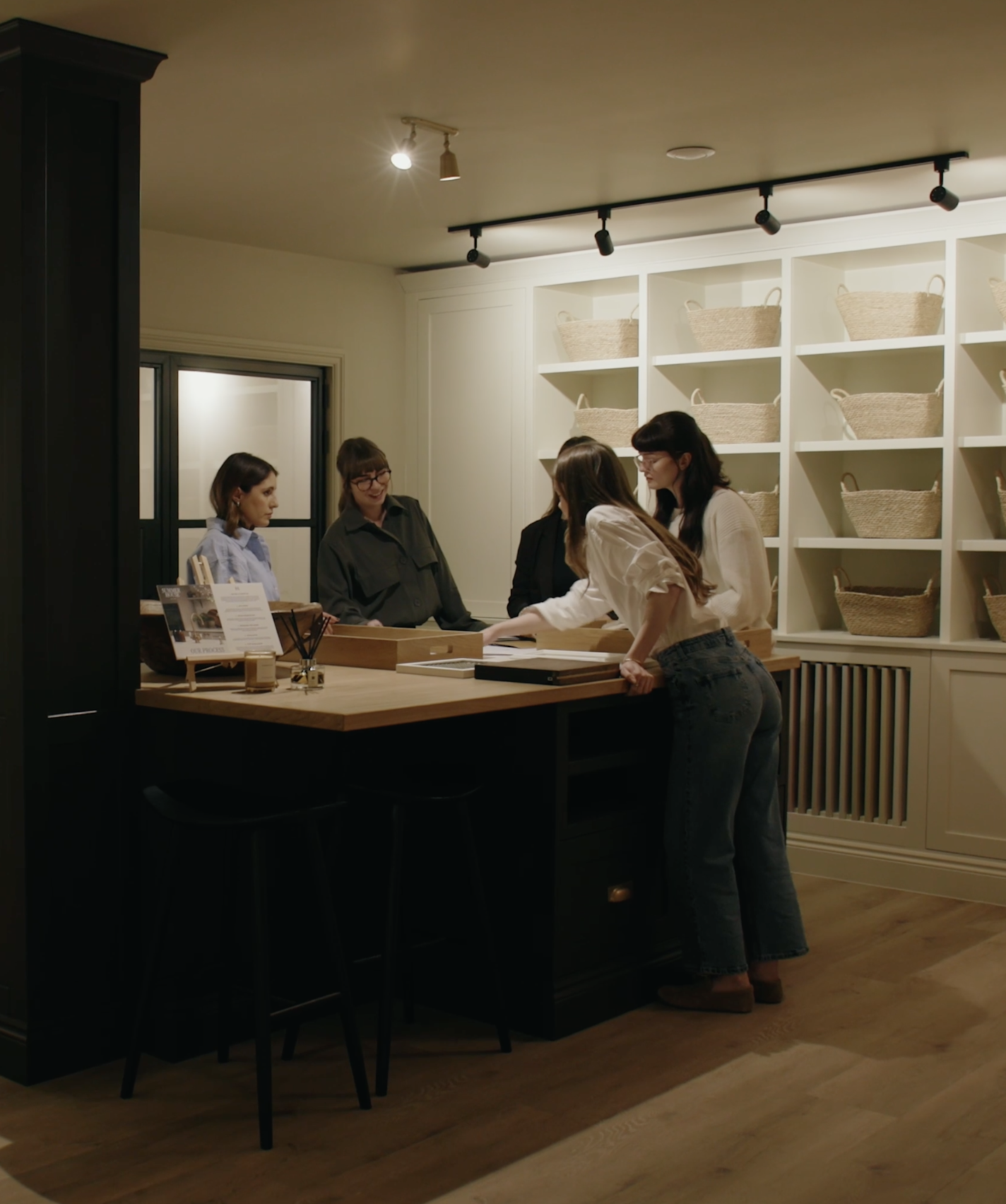 Four women gathered around a wood-topped table in a room with white shelving filled with baskets on the wall. They appear to be discussing or working on something.
