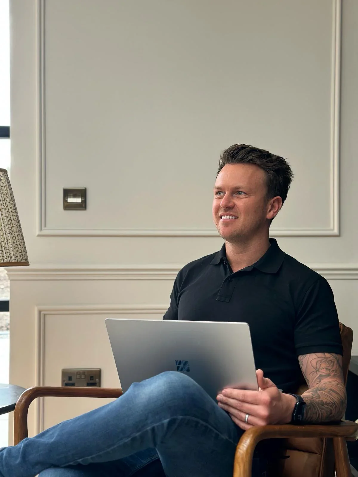 A man with tattoos on his arm sitting in a wooden chair, holding a laptop, smiling thoughtfully in a room with cream-colored walls and a window.