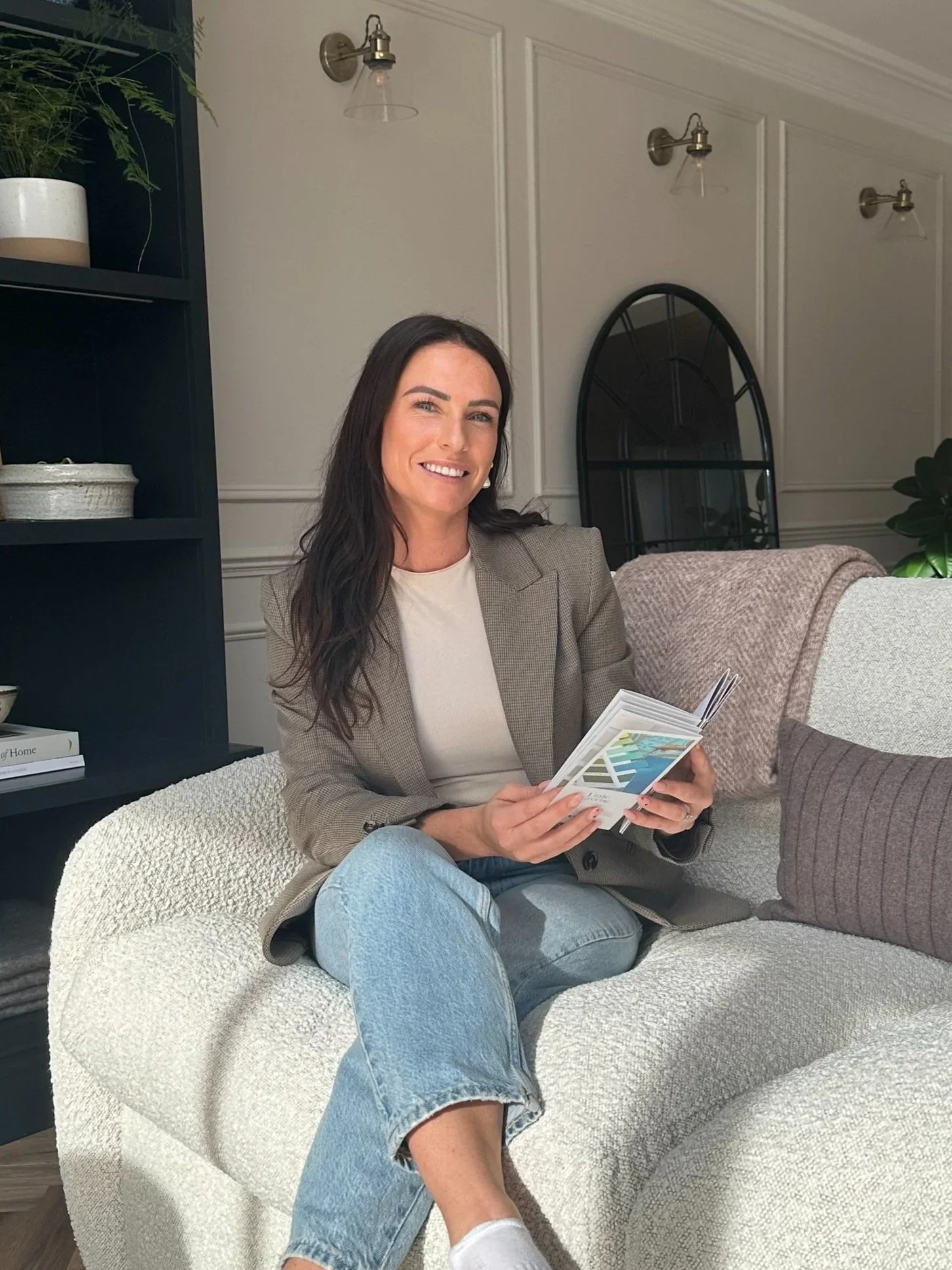 A woman with dark hair, wearing a beige blazer and jeans, sitting on a cream-colored textured couch and holding a color palette or swatches in her hands, smiling.