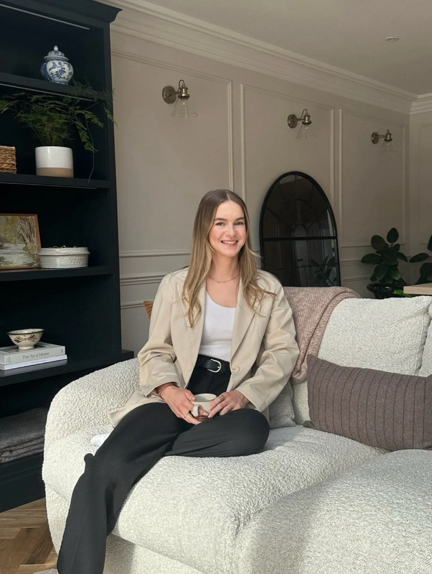 A woman sitting on a beige sofa with a white coffee mug in her hand, in a cozy living room, smiling at the camera. She has long blonde hair, wears a light-colored blazer and black pants. The background features a black bookshelf with decorative items, a black framed window, and a large potted plant.