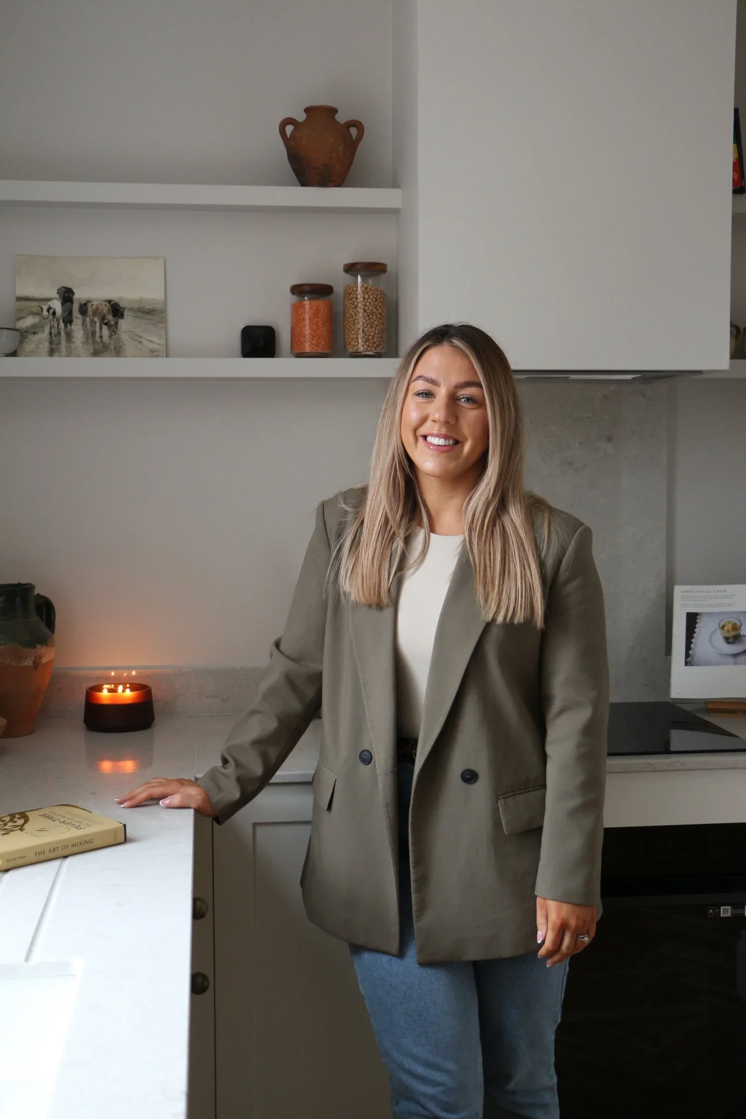 A woman with long blonde hair smiling and standing in a modern kitchen, wearing a beige blazer and blue jeans. She has one hand resting on a white countertop, which has a book titled 'The Art of Mixing,' a lit candle, and a vase. Behind her are white shelves with decorative items including a painting of cows, jars of spices, a black speaker, and a large brown vase.