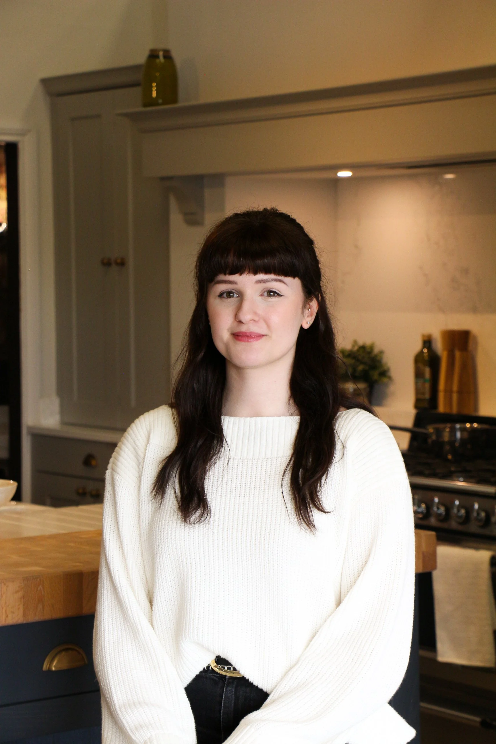 A woman with dark brown hair and bangs, wearing a white sweater, standing in a kitchen.