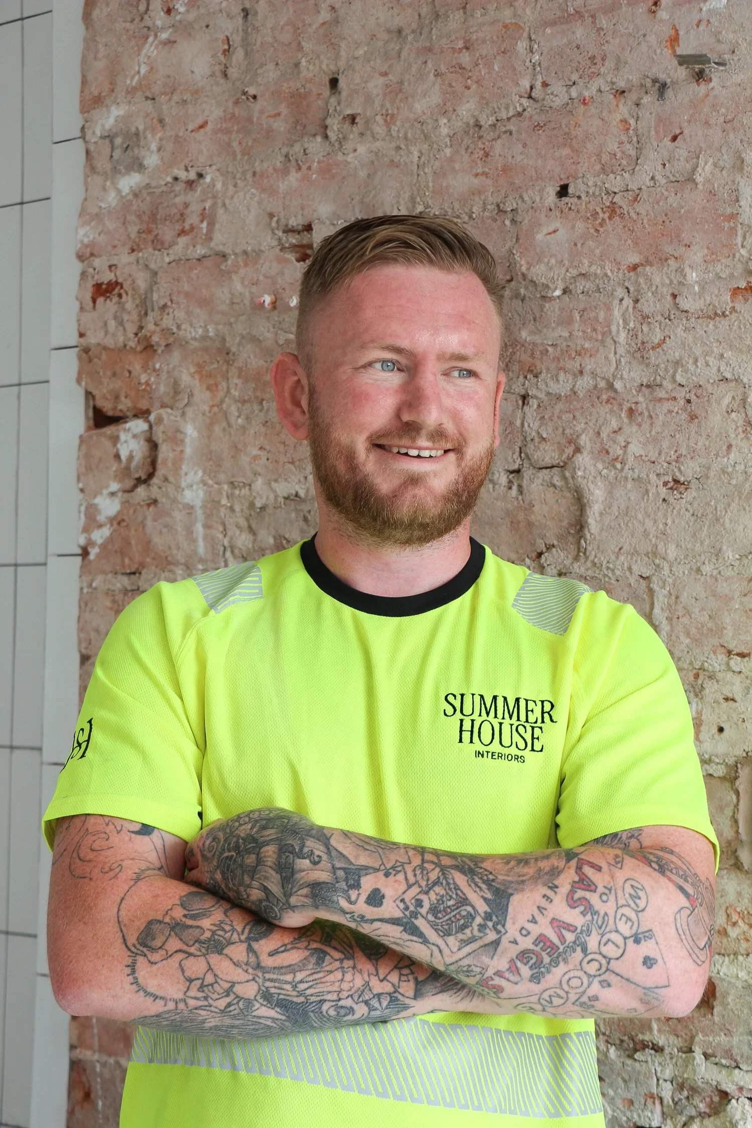 A smiling man with short, styled hair and a beard, wearing a bright yellow shirt with black lettering that reads 'SUMMER HOUSE INTERIORS.' He has extensive tattoos on his arms and is standing against a brick wall, with his arms crossed.