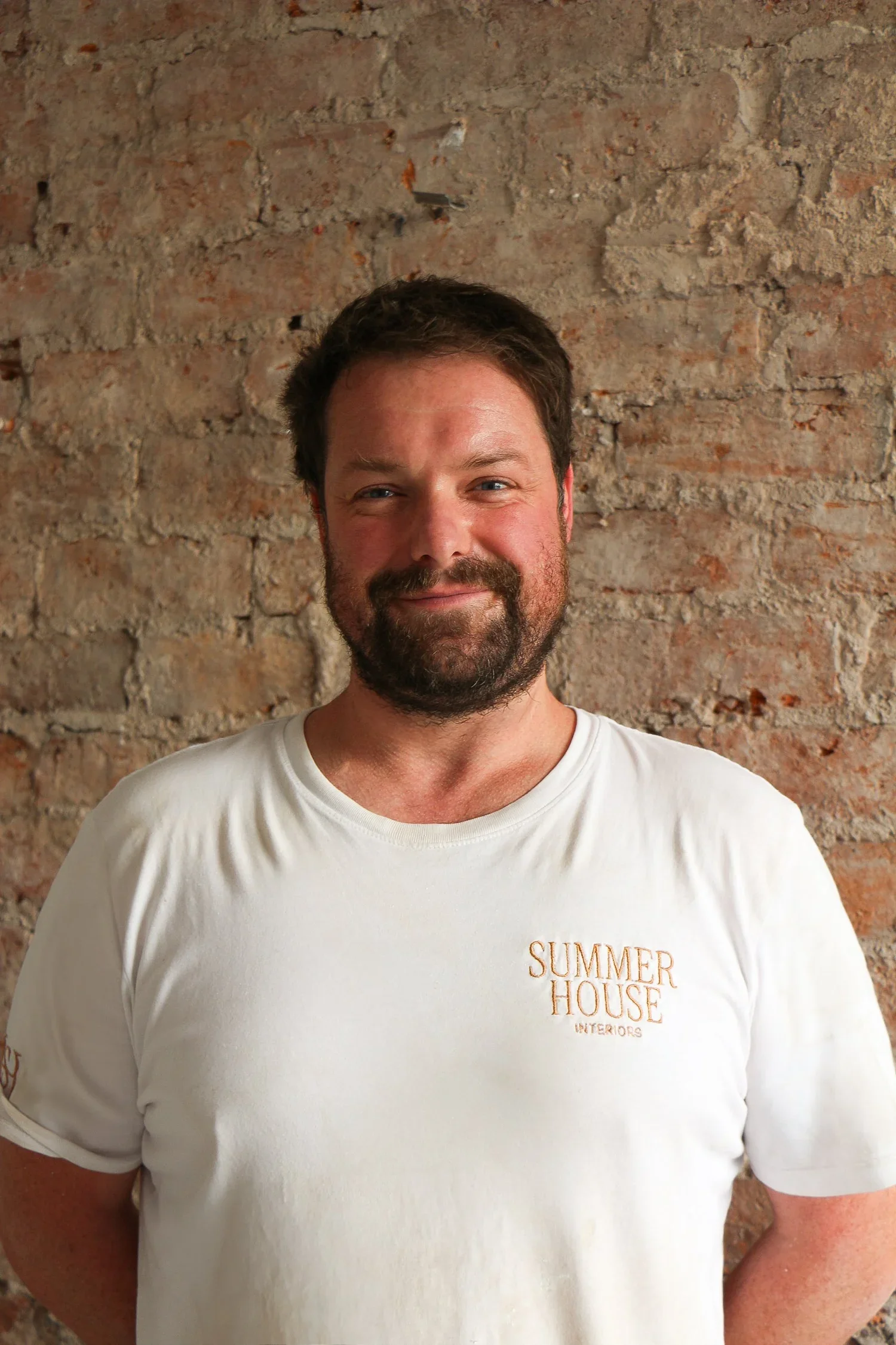 Close-up of a man smiling, wearing a white t-shirt with gold lettering that reads 'Summer House Interiors,' standing in front of a brick wall.