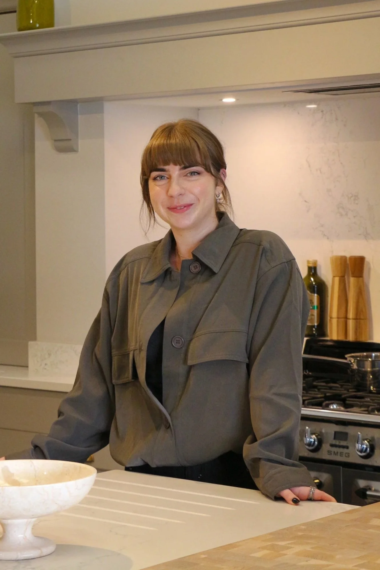 A woman with brown hair and bangs, wearing a gray jacket, stands in a kitchen with a stove and kitchen utensils in the background. She is smiling and leaning on a white kitchen counter with a large white bowl in front of her.