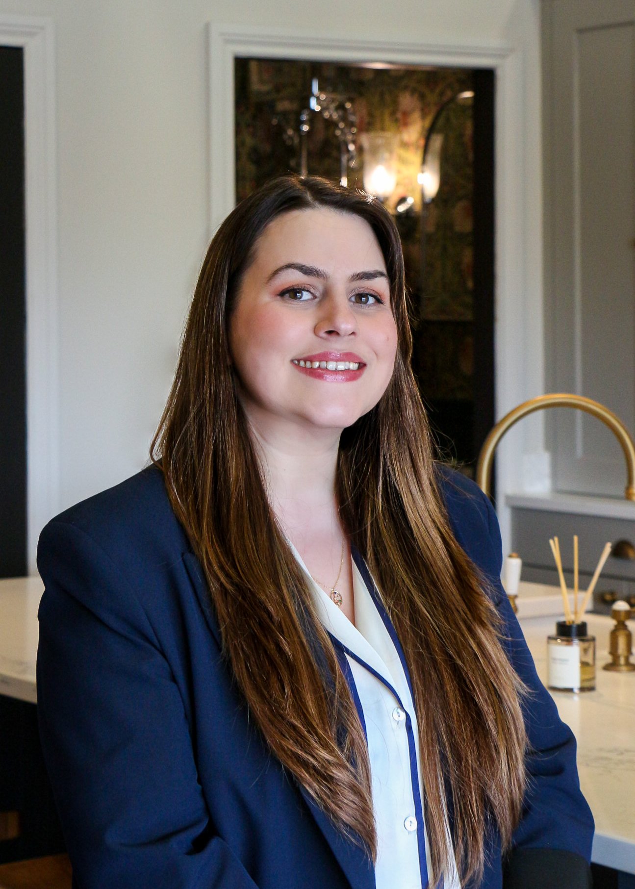 A woman with long brown hair, wearing a navy blazer and white blouse, smiling in a modern kitchen with a white countertop, gold sink faucet, and decorative items in the background.