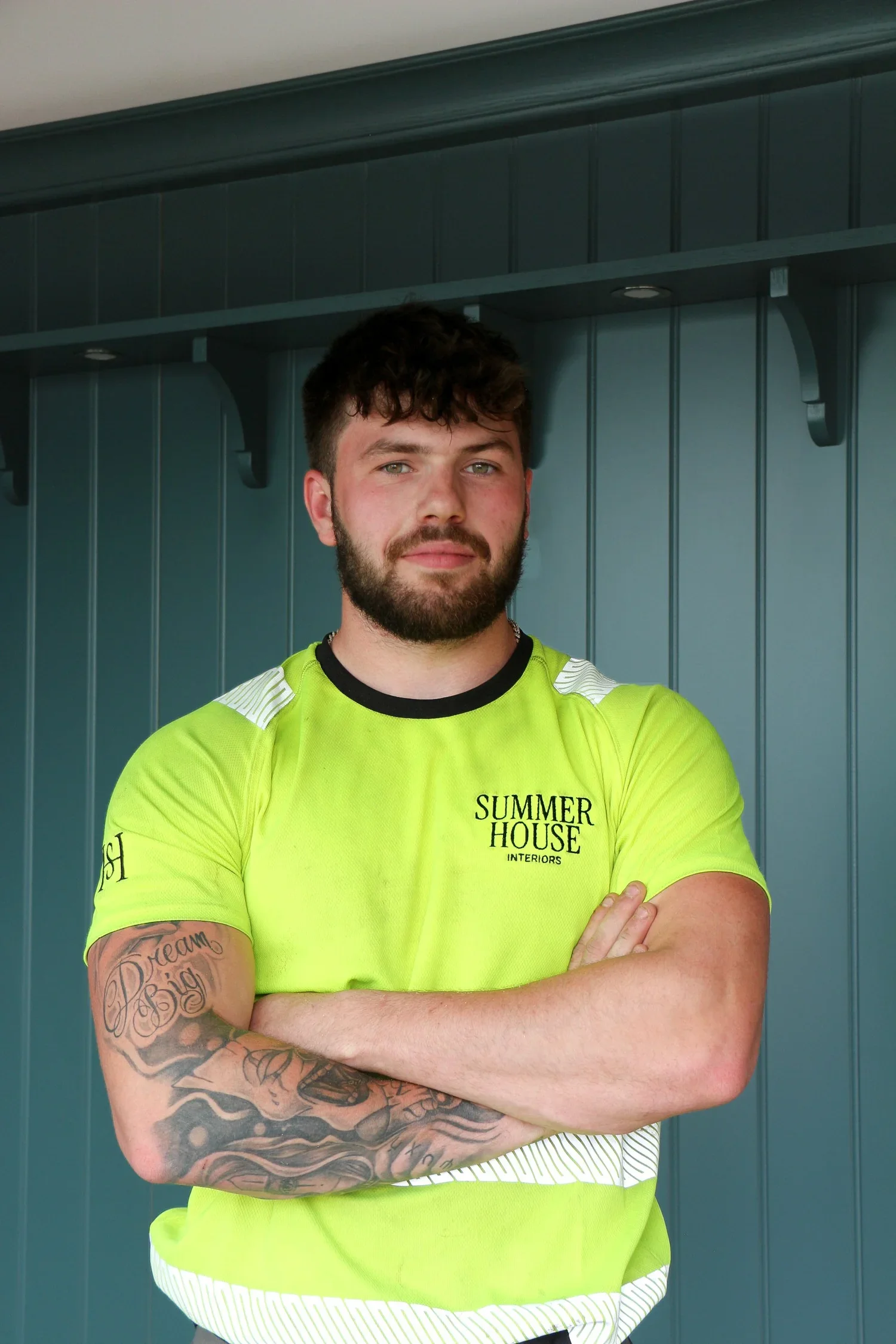 A young man with a beard and tattoos on his right arm, wearing a neon yellow sports shirt, standing in front of a blue wall with a shelf above his head. His arms are crossed.