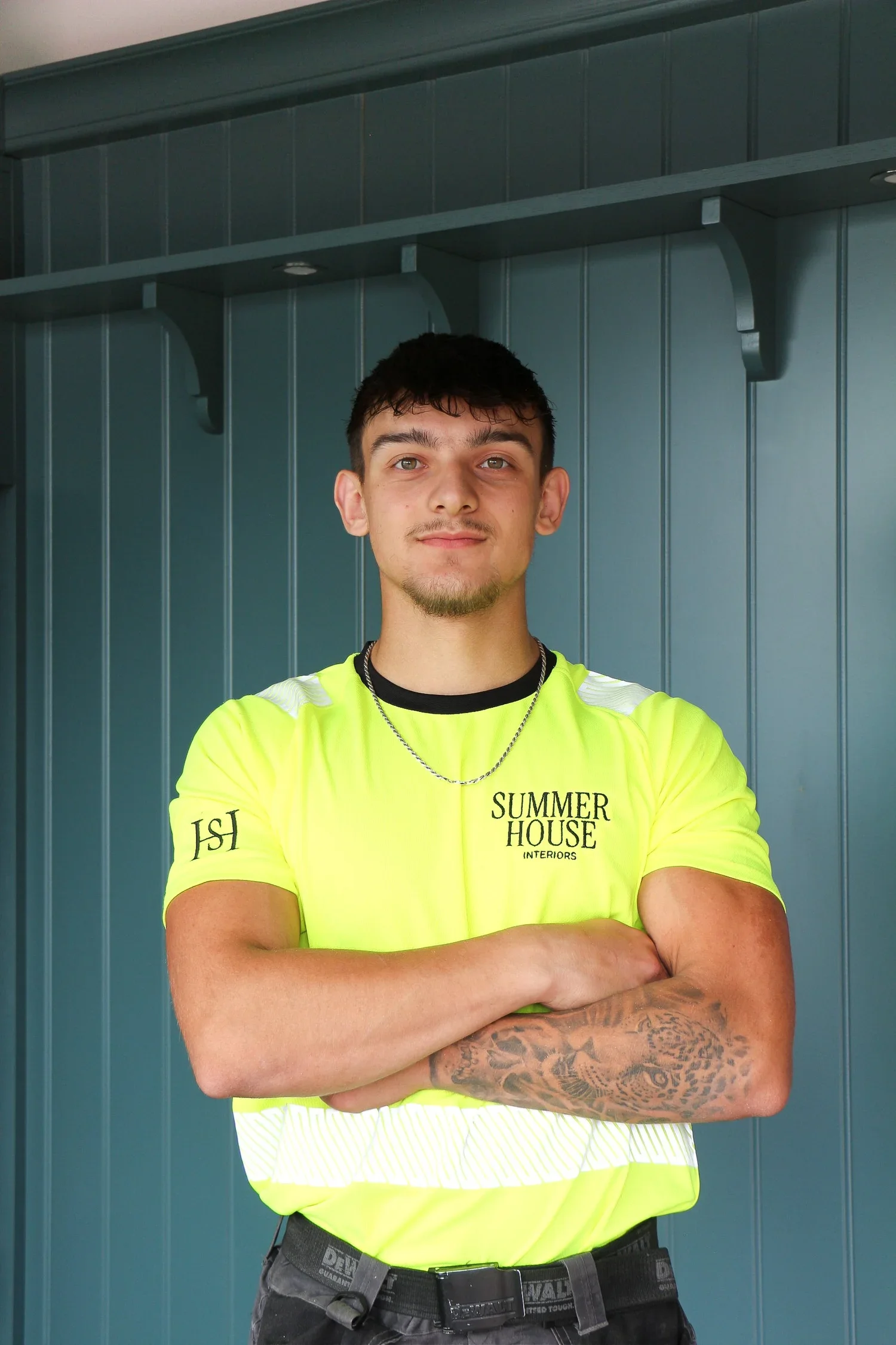 Young man with short dark hair, tattoos on his right arm, wearing a bright yellow high-visibility shirt with 'SUMMER HOUSE INTERIORS' written on it, standing with arms crossed in front of a teal wall with a shelf above.