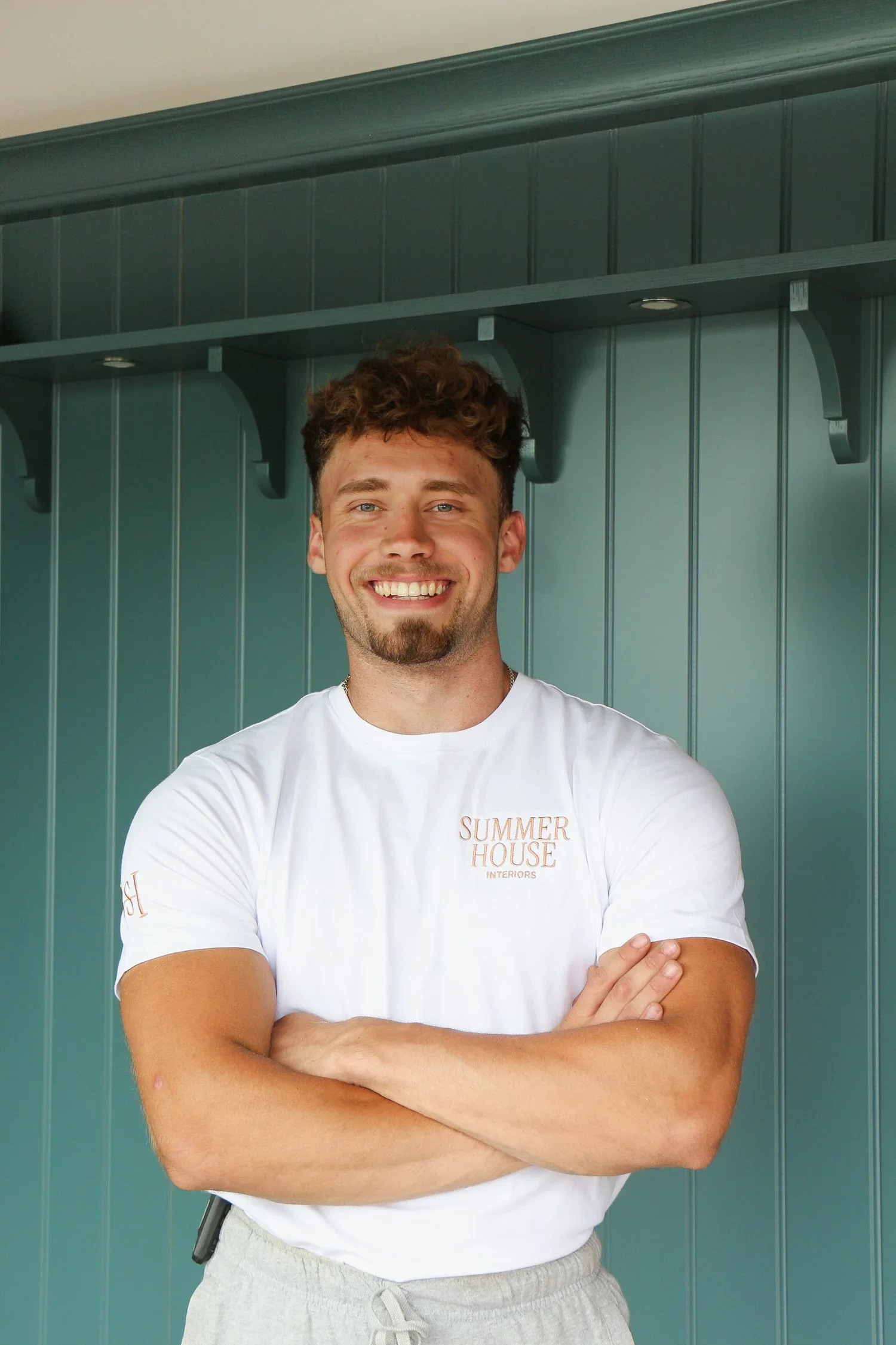 smiling young man with curly hair, crossed arms, wearing a white T-shirt with 'SUMMER HOUSE INTERIORS' logo, standing in front of a green wooden wall.
