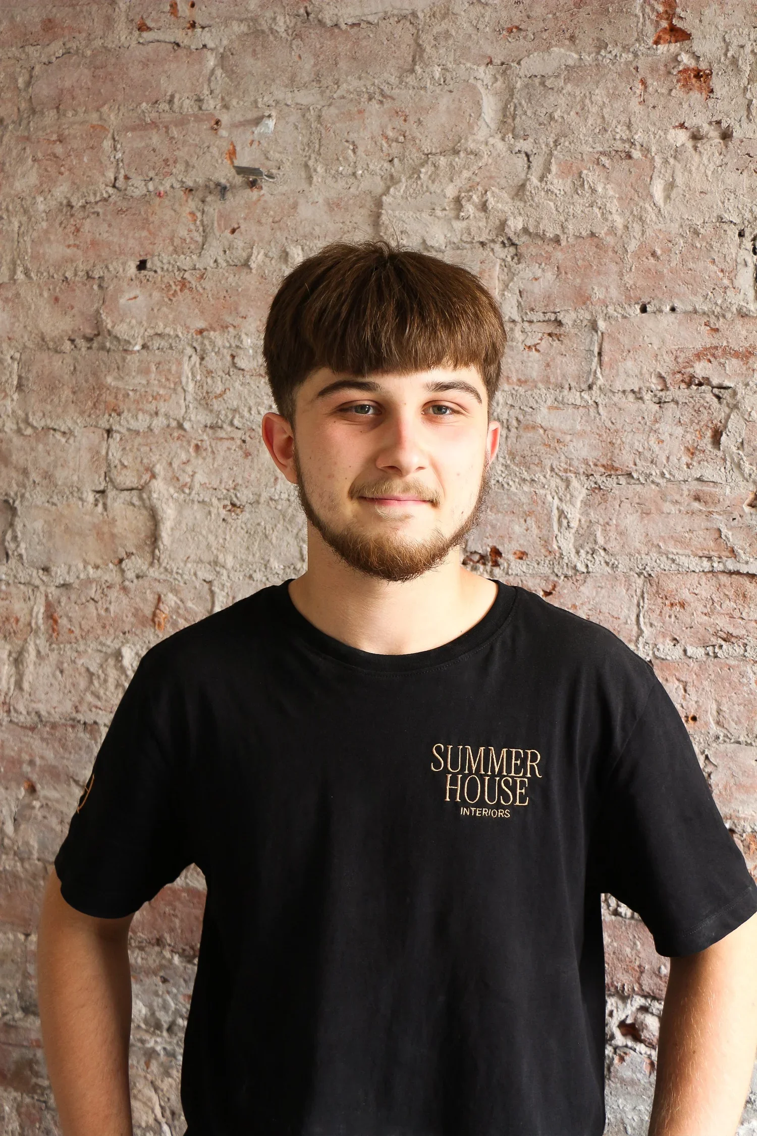 A young man with light skin and brown hair standing against a brick wall, wearing a black t-shirt that says 'Summer House Interiors'.