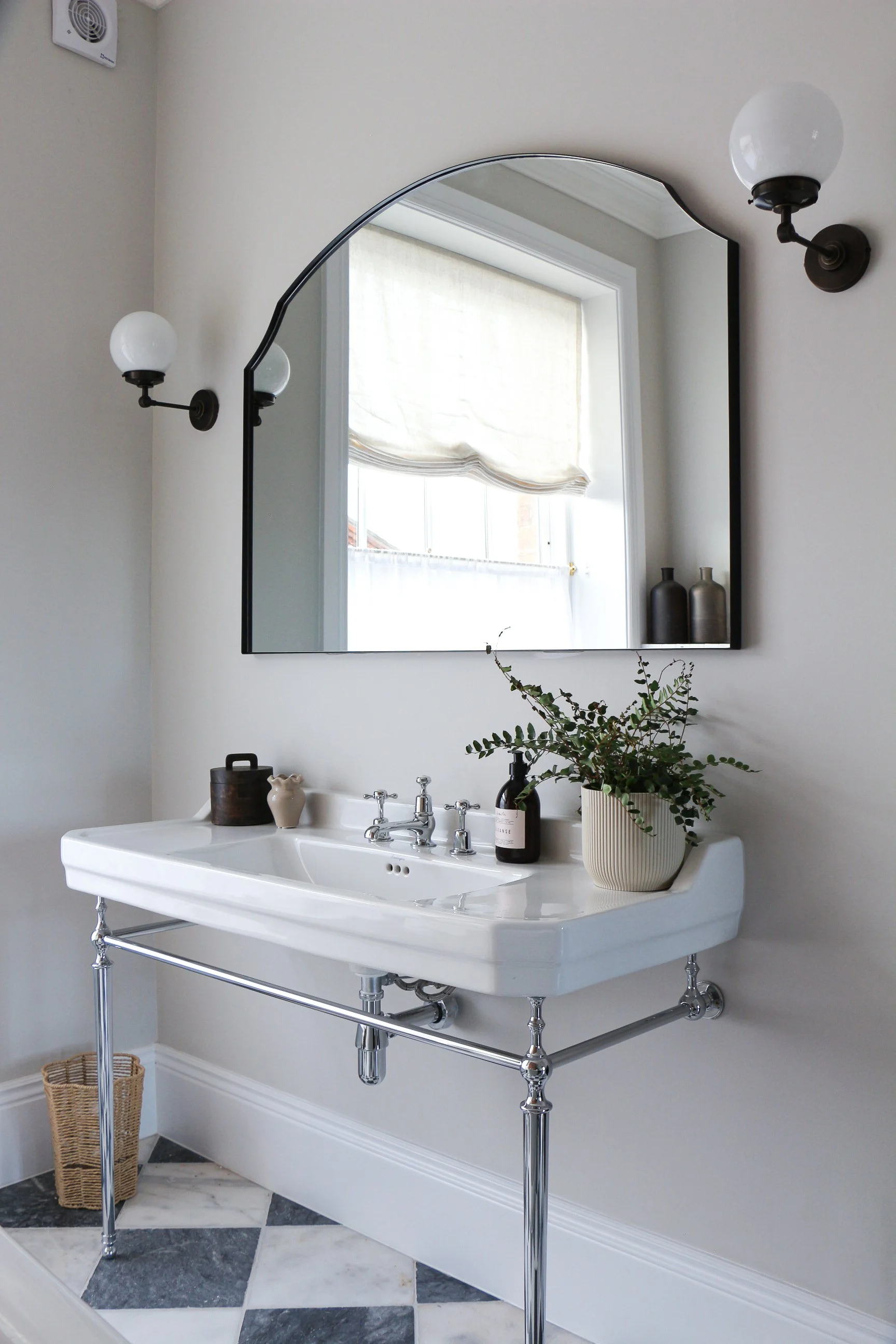 A vintage white sink with chrome legs in a bathroom, decorated with a potted plant, soap, and vases. A mirror, window, and wall sconce are visible behind the sink. The floor has black, white, and gray tiles.