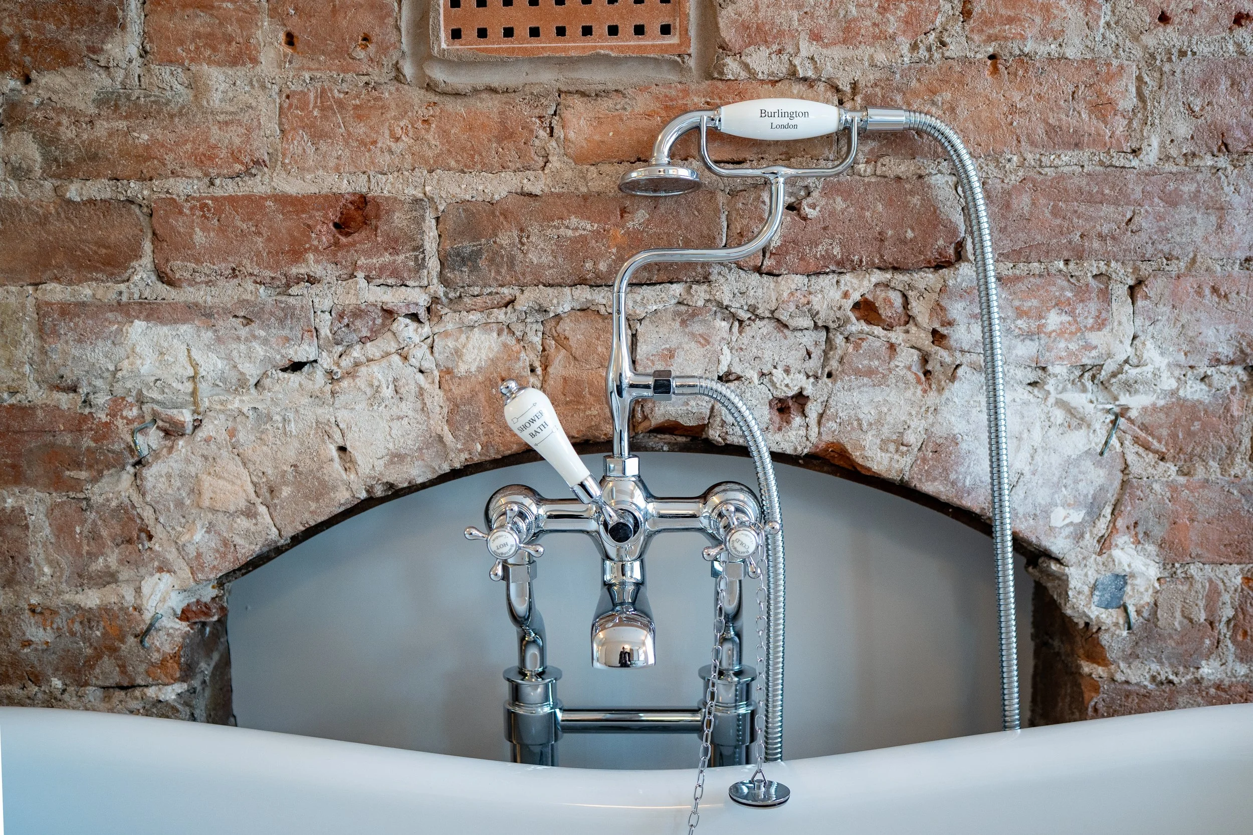 Close-up of a vintage-style bathroom faucet on a white bathtub, set against a rustic exposed brick wall.