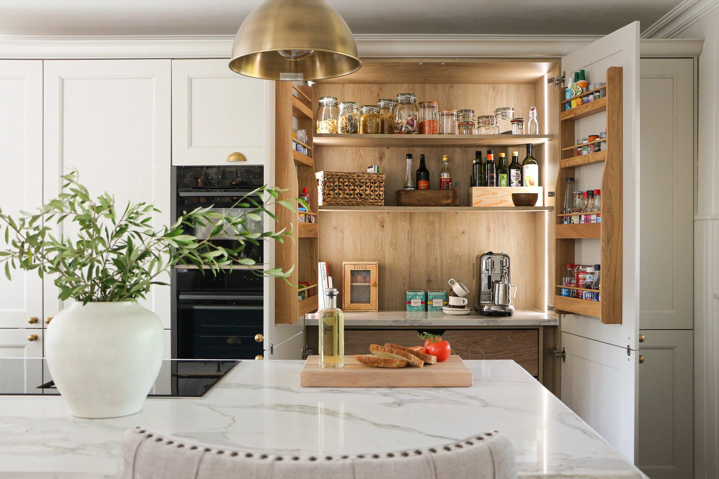 A modern kitchen with white cabinets and open wooden shelving containing jars and bottles. A marble kitchen island in the foreground has a white vase with green foliage, a bottle of olive oil, sliced bread, a tomato, and a cloth. There is a built-in stove and oven, with a small wooden wine rack and various kitchen items on the open shelves.