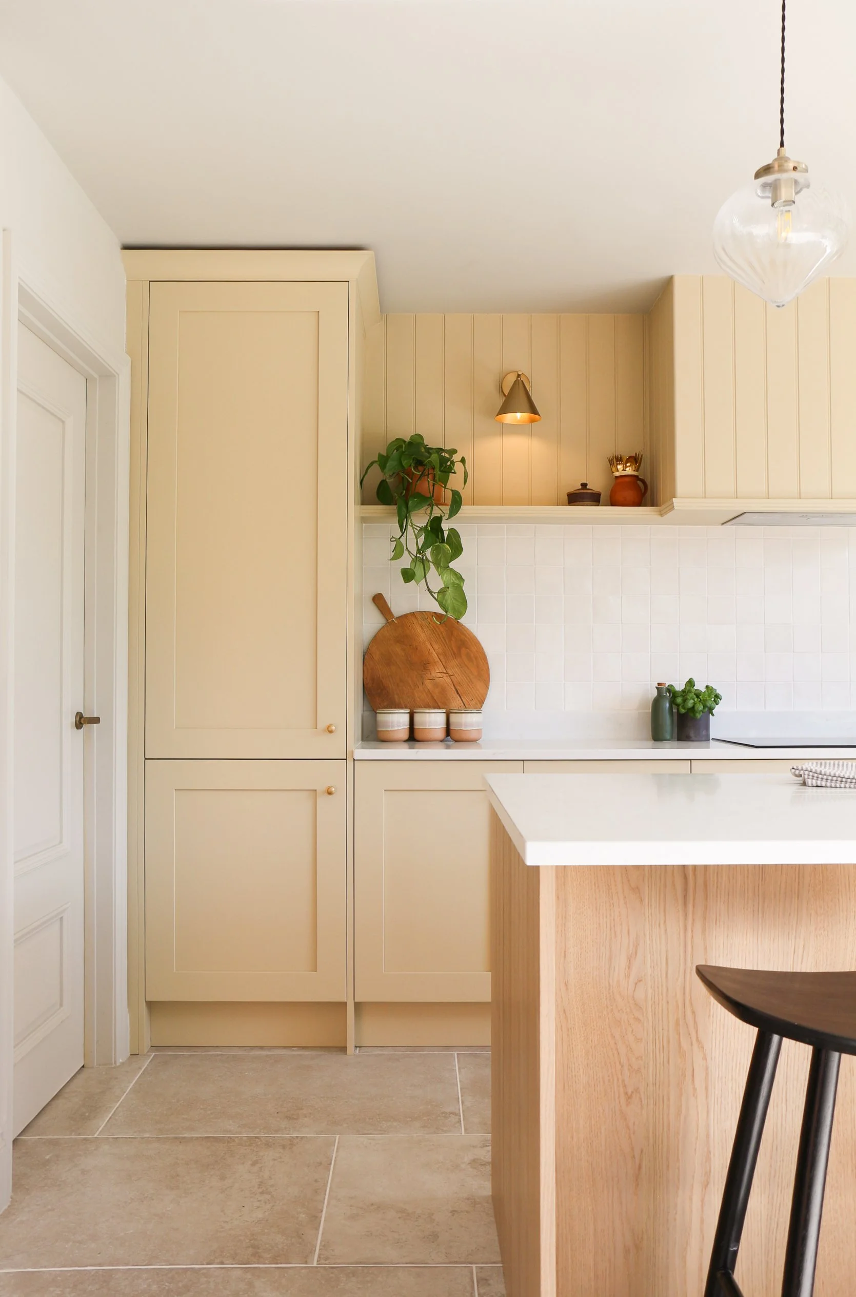 A kitchen section with beige cabinets, a white countertop, a potted plant, a wooden cutting board, three jars, and a wall-mounted light fixture. Part of a black chair is visible in the foreground.