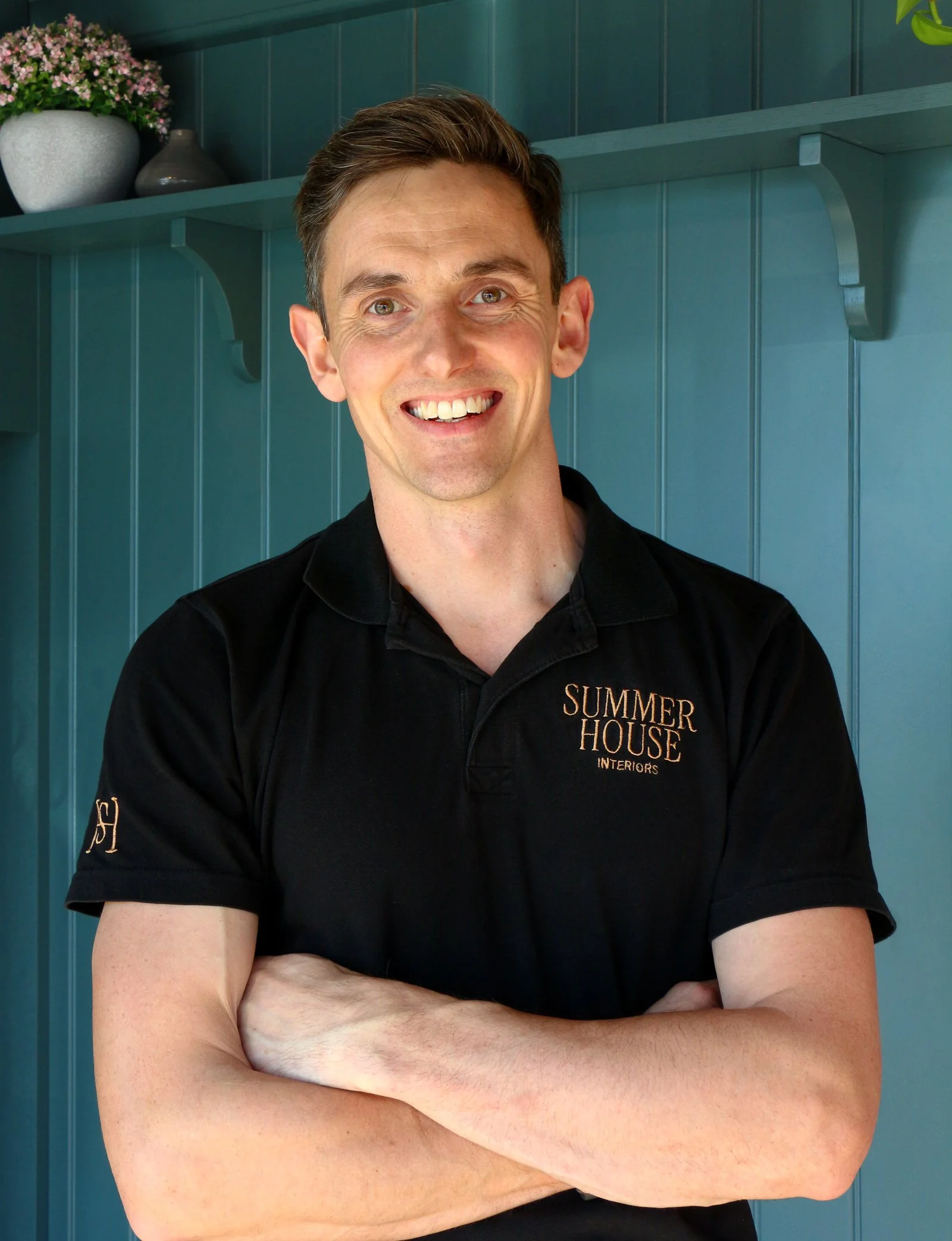 A man with short brown hair smiling, crossing his arms, wearing a black polo shirt with embroidered gold text reading 'SUMMER HOUSE INTERIORS', standing in front of a teal-blue wooden wall with a floating shelf that has a potted pink flowering plant and two decorative vases.