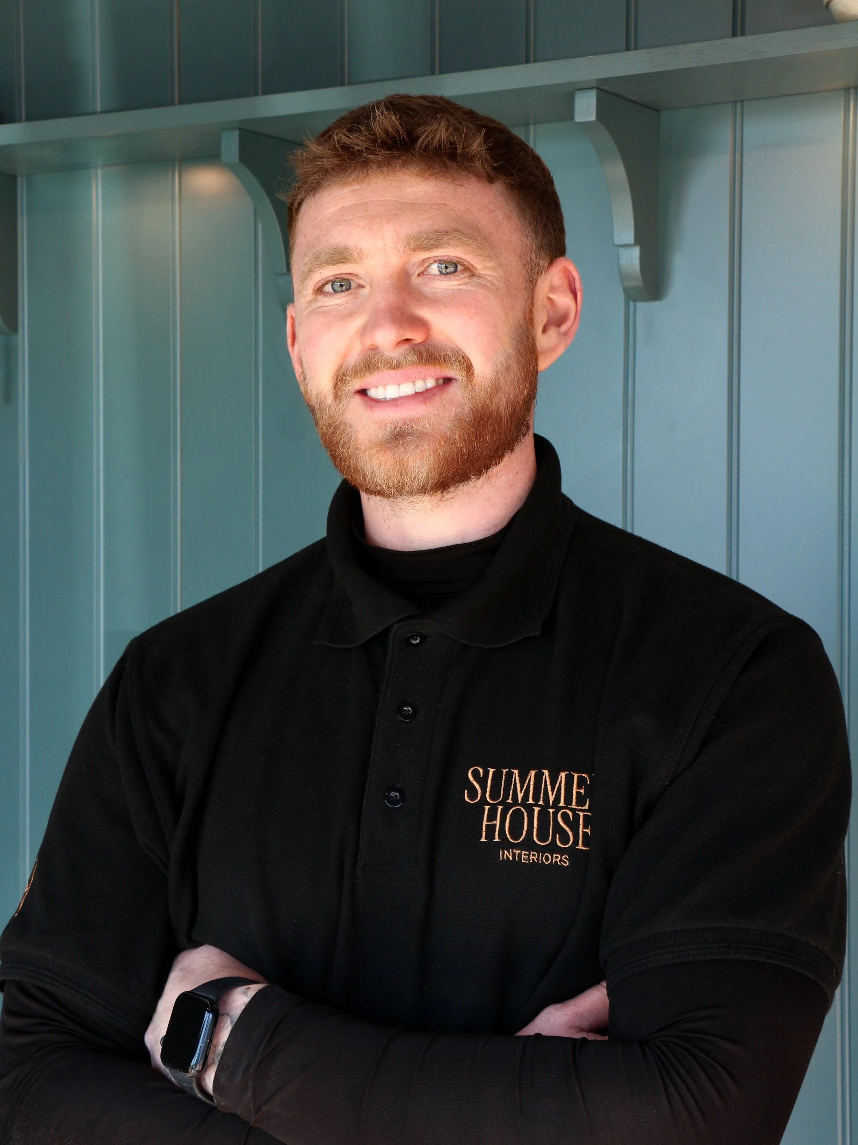 A man with short brown hair, a beard, and blue eyes is smiling and standing with arms crossed. He is wearing a black shirt with gold lettering that reads "SUMME HOUSE INTERIORS". The background features teal-colored paneling.