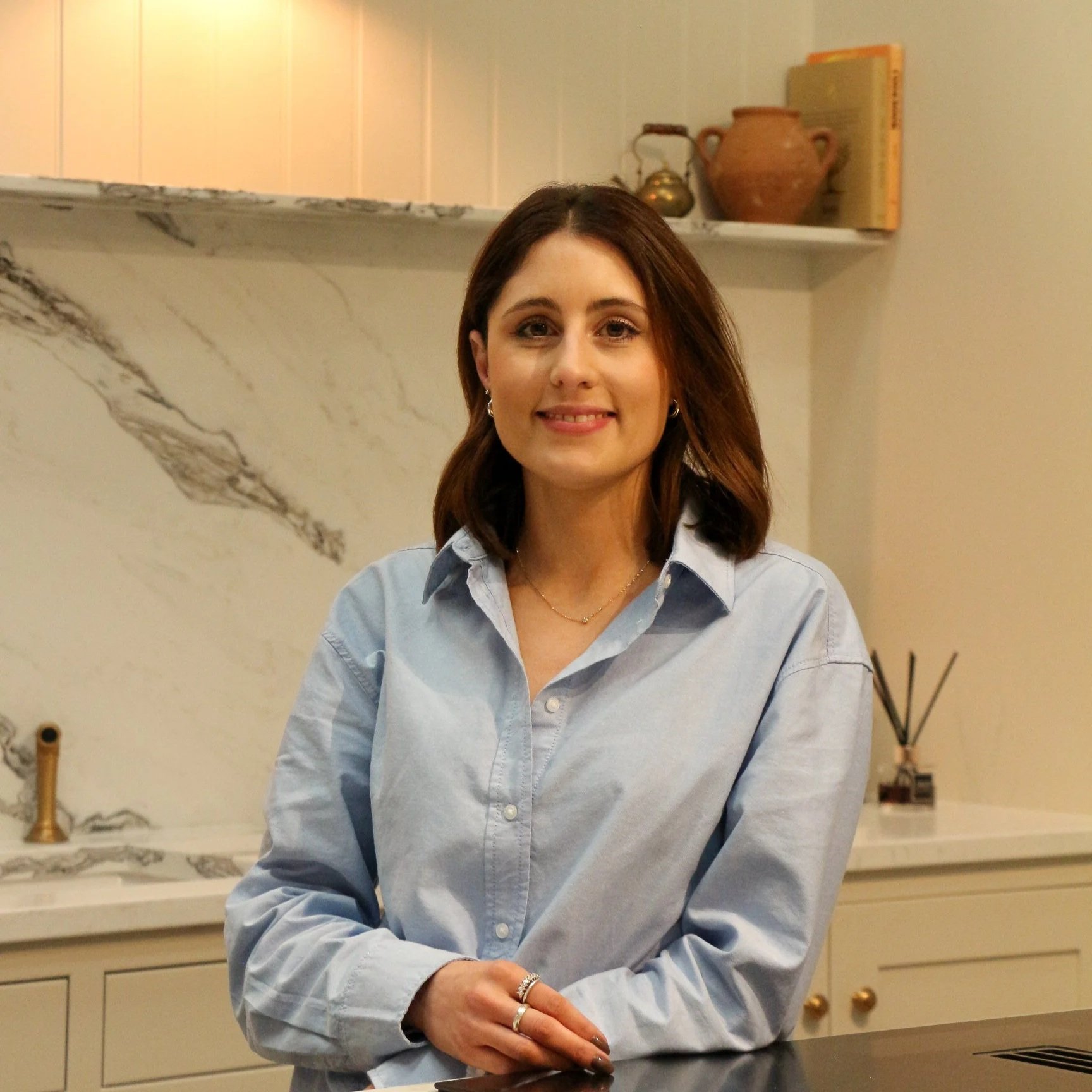 A young woman with shoulder-length brown hair, wearing a light blue button-up shirt, smiling while sitting at a kitchen counter. The background features a marble backsplash, a shelve with decorative items including a clay vase, a book, and a brass kettle.