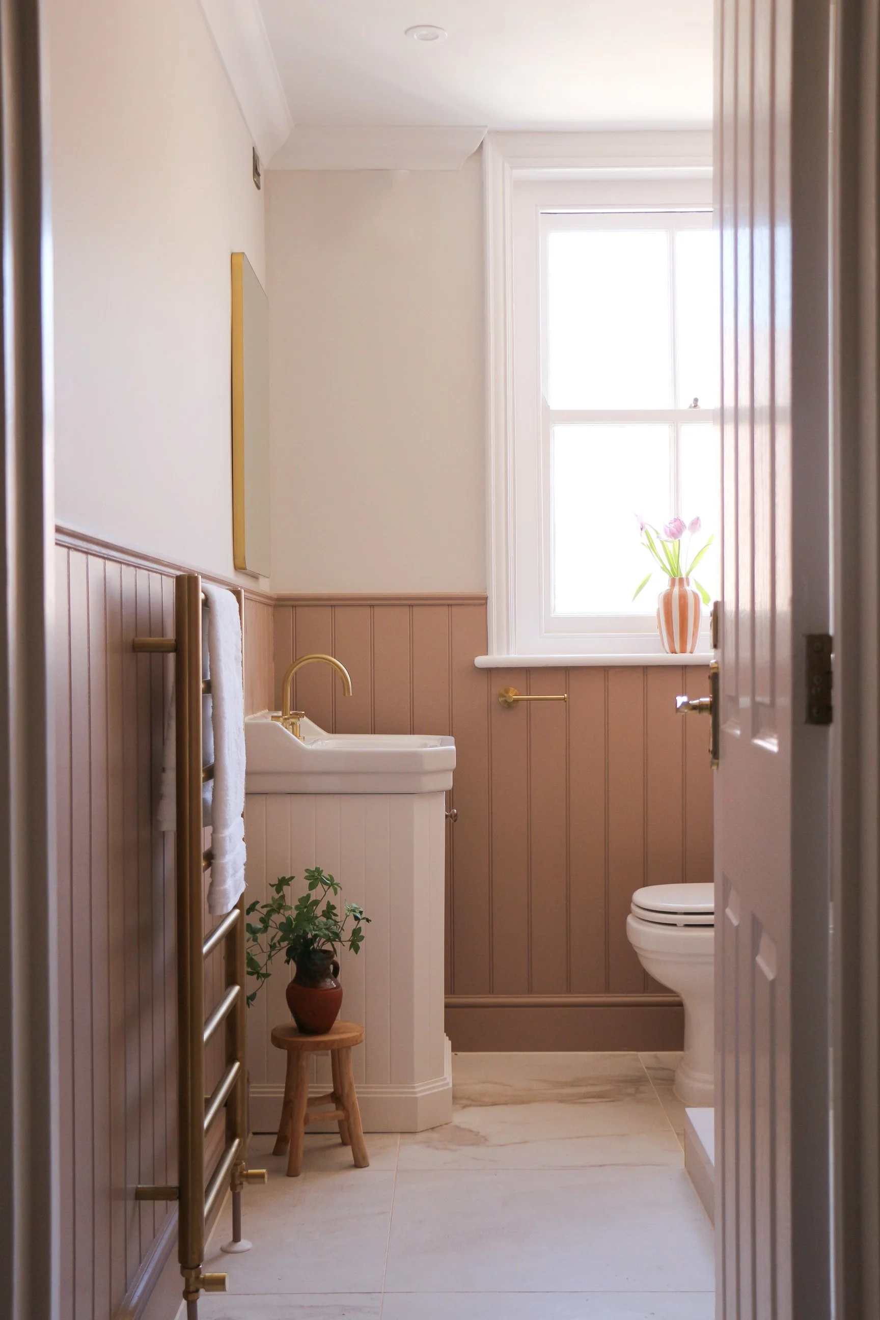 A small bathroom with a window, a vessel sink, a toilet, a towel rack, a small potted plant, and a vase with pink flowers.